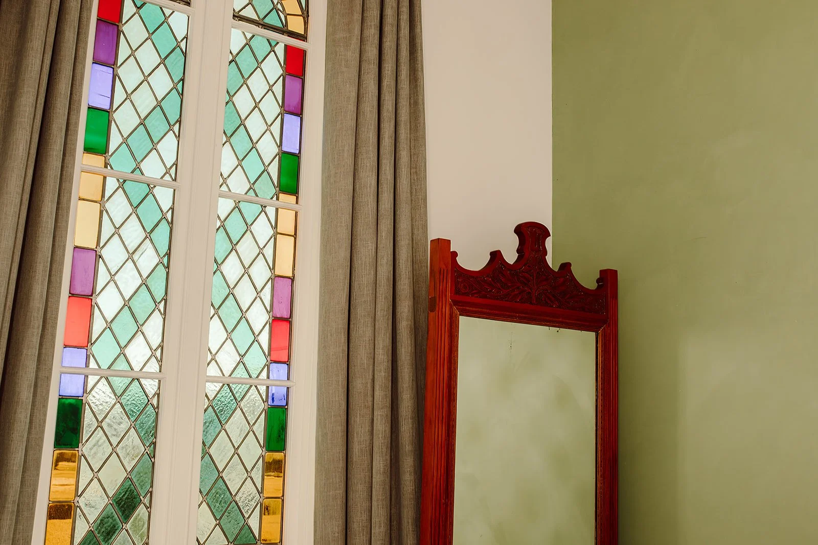 A stained glass window with colorful geometric patterns next to a silver curtain, and a partially visible red carved wooden mirror frame against a pale green wall.