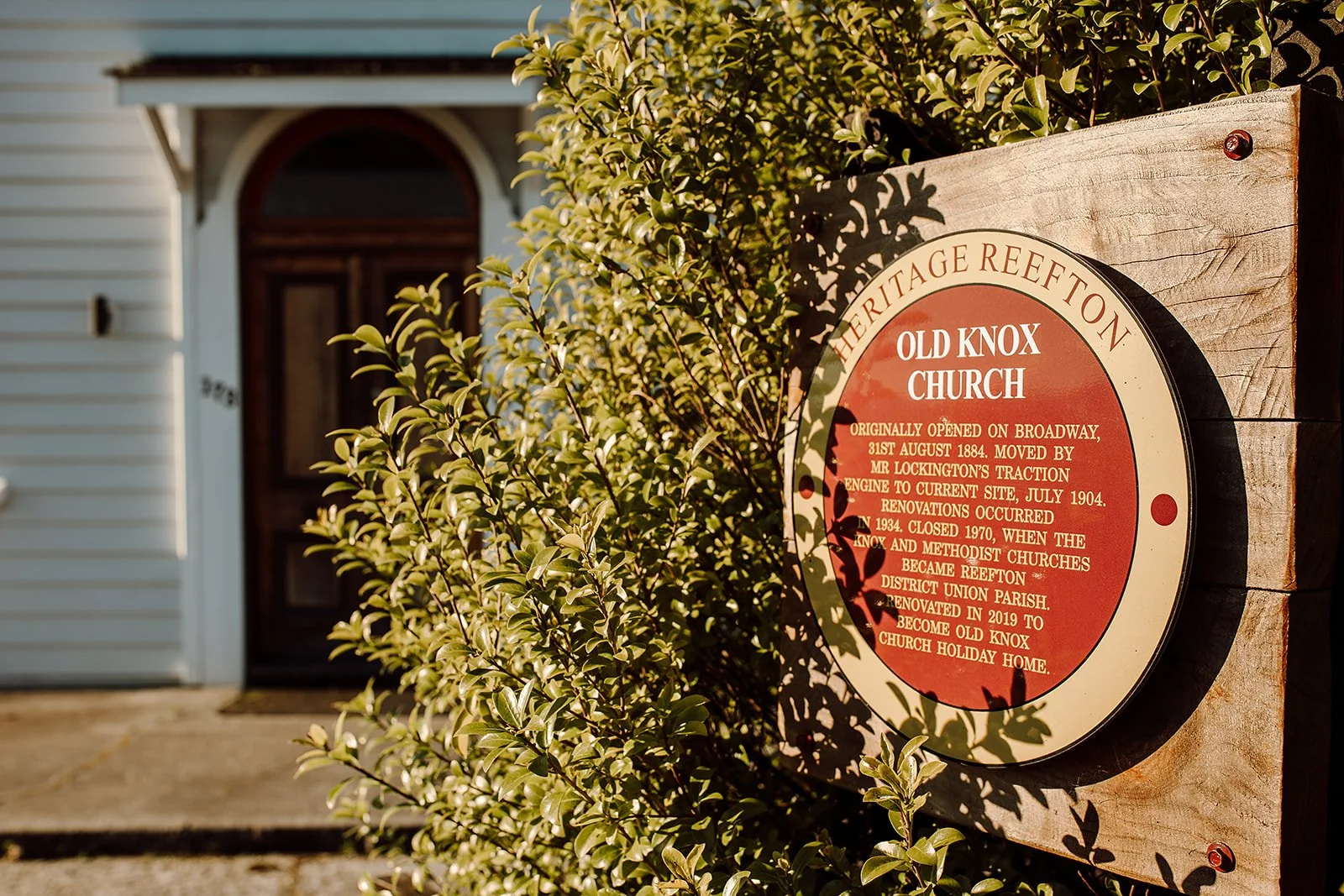 A historical plaque on a wooden board provides information about Old Knox Church, located near a leafy bush in front of a house with a dark wooden door. The plaque details the church's history, including its original opening, relocations, renovations