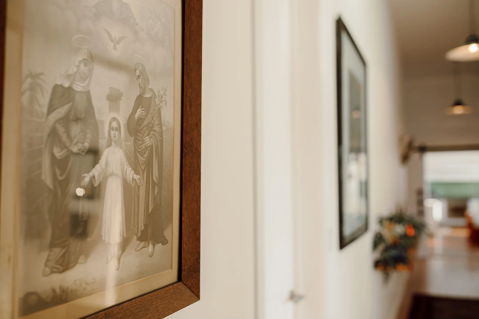 Close-up of religious artwork framed on a white wall in a hallway, with photos and a flower arrangement visible further down the corridor.