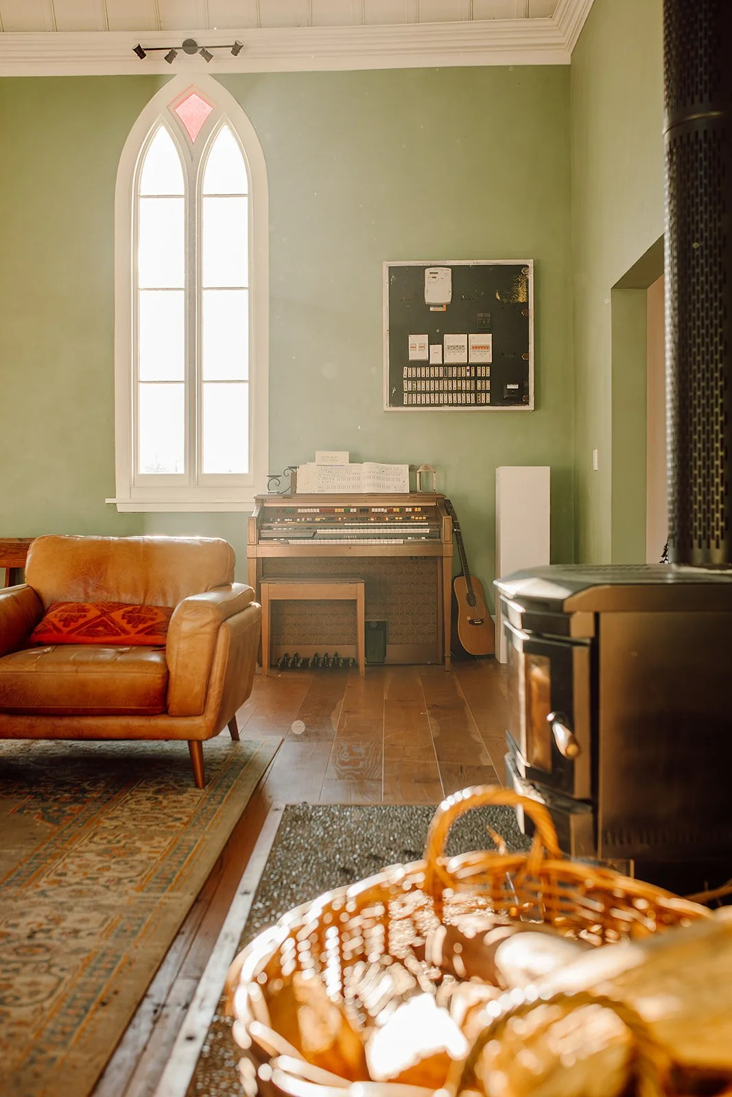 Interior of a cozy room with a large arched window, a vintage leather sofa with a red pillow, an old upright piano with sheet music on top, an acoustic guitar leaning against the piano, a wood-burning stove, a basket with firewood, and a patterned ru