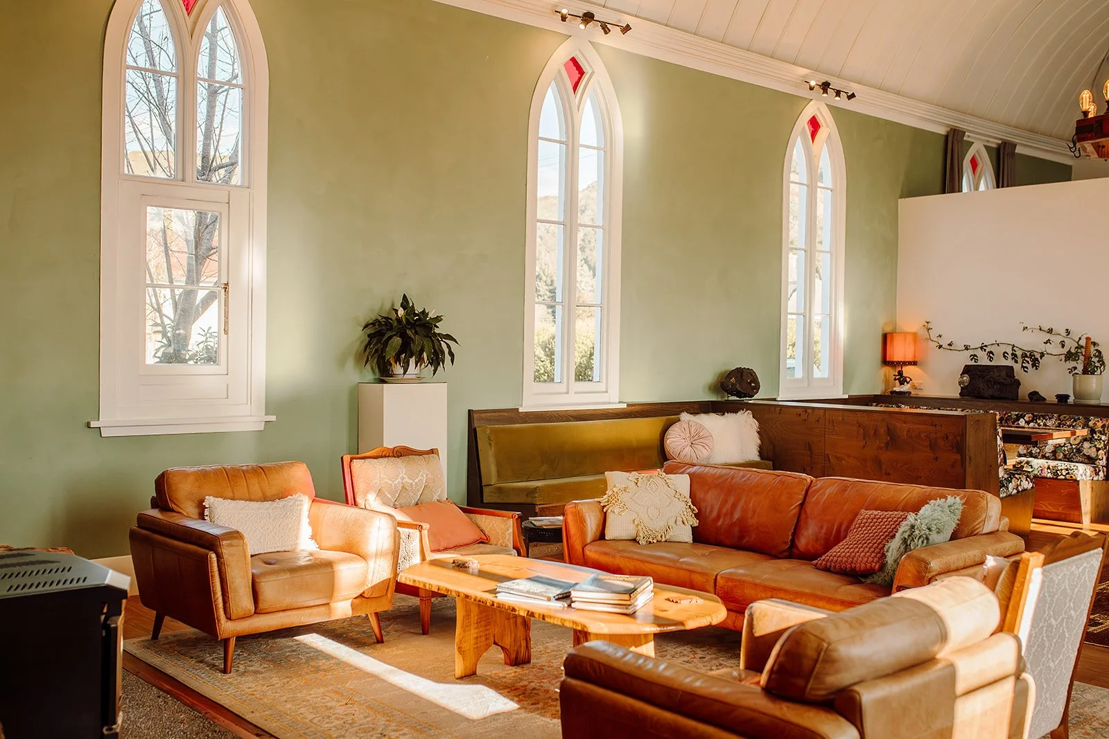 A cozy living room with green walls, three tall arched windows, and vintage furniture including a leather sofa and armchairs, a wooden coffee table, a plant on a white stand, and warm lighting.