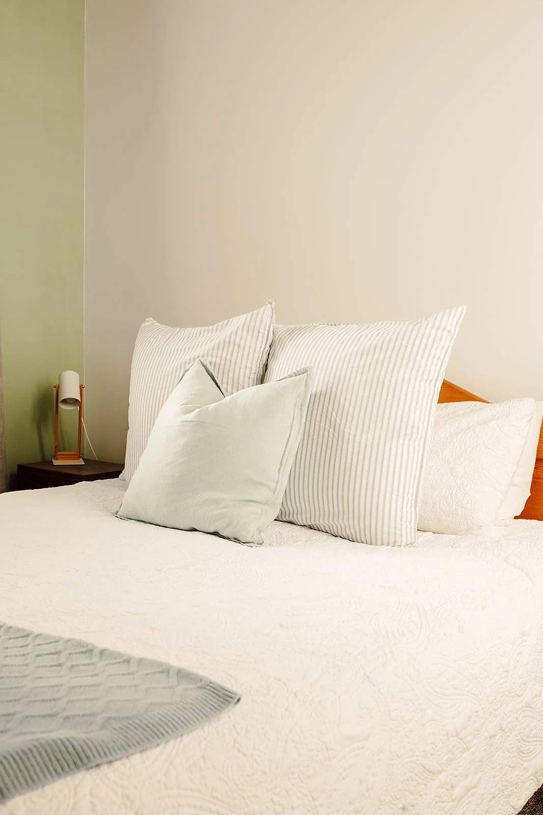 A neatly made bed with white and striped pillows on a bed with a textured white bedspread, next to a nightstand with a small lamp.