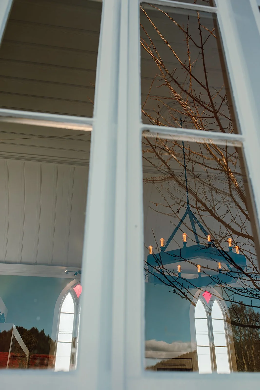 Reflection of a chandelier with candles, tree branches, and arched windows in a glass door