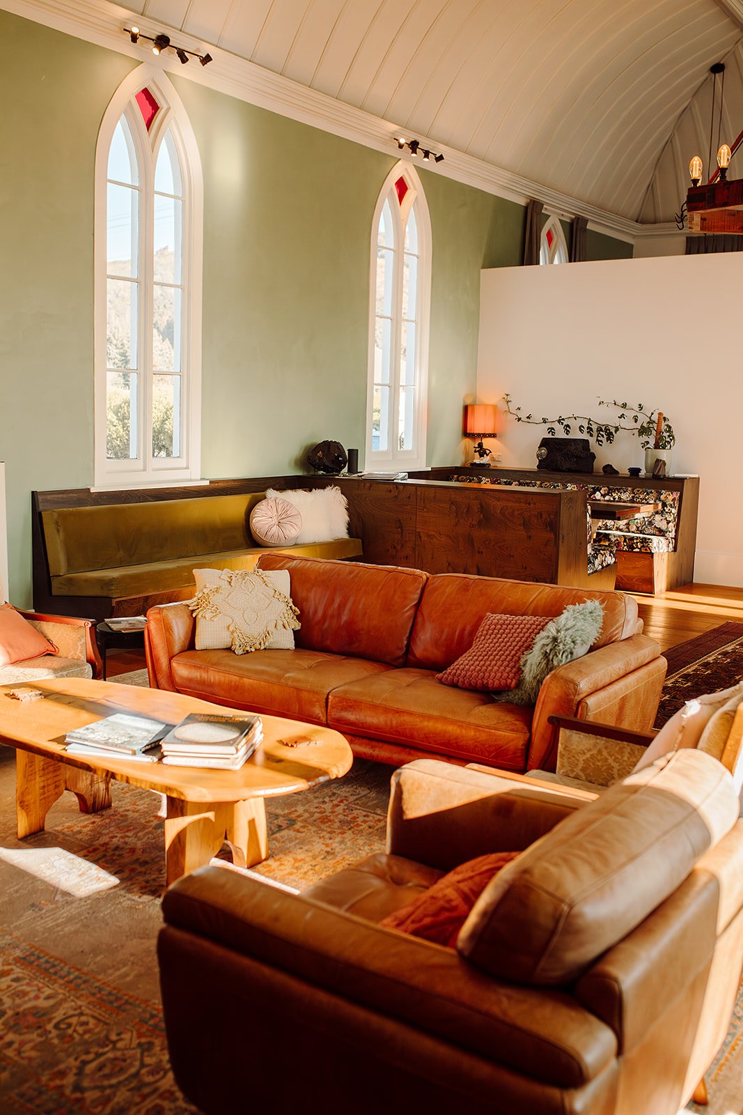 Living room with green walls, three tall arched windows with red accents, a leather sofa, a wooden coffee table, and a corner with a built-in wooden bench and a bar area with a lamp and decorative plants.