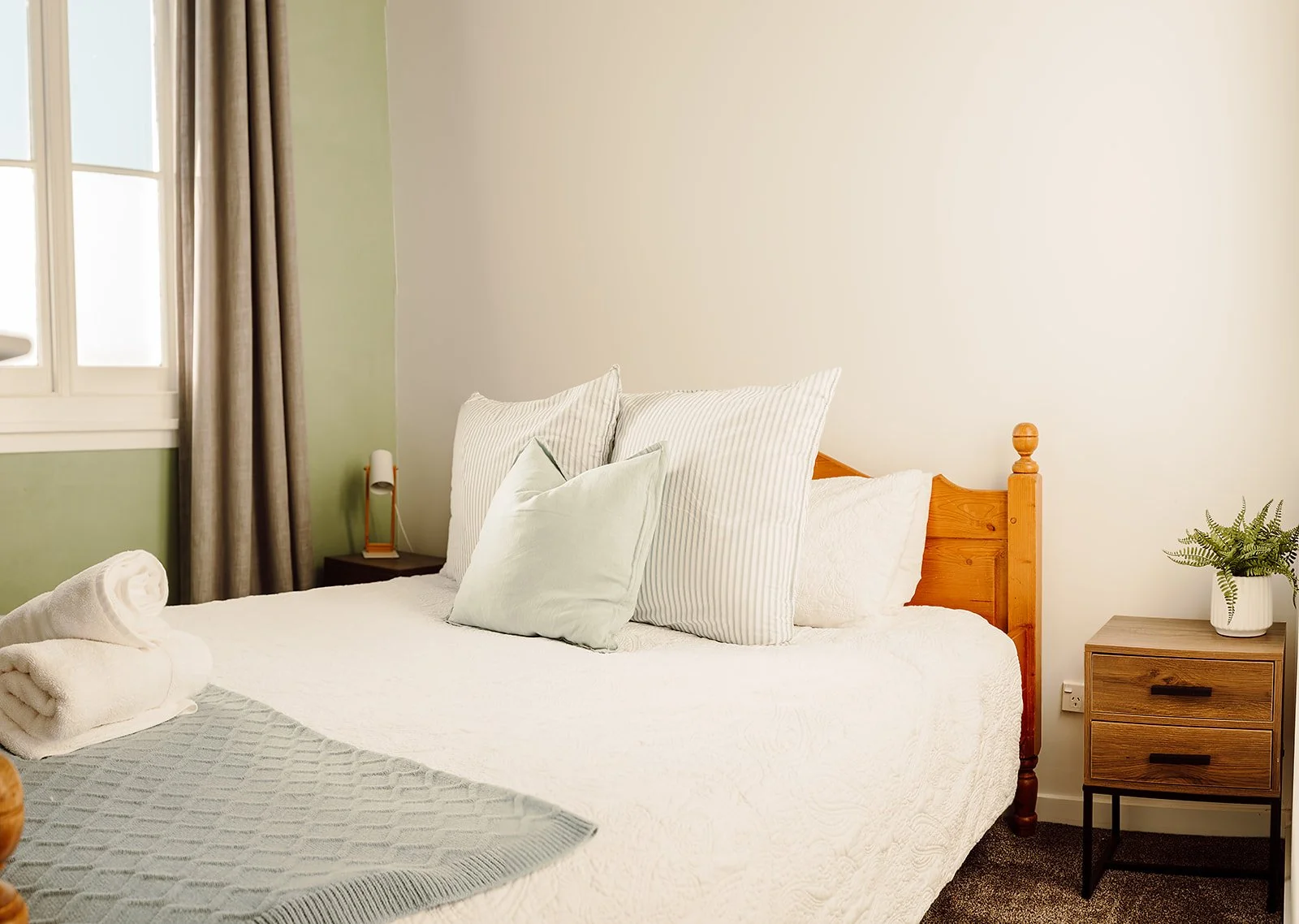 A neatly made bed with white linens and multiple pillows in a bedroom. There is a small wooden nightstand with a potted plant on it on the right side, and a second nightstand with a lamp on the left. The window has beige curtains, and the room has li