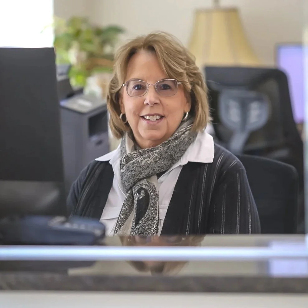 Smiling woman with glasses and scarf at her office desk.