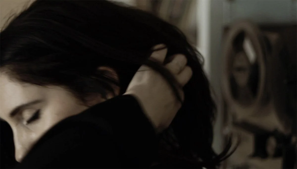 Soft-focus portrait of a person with eyes closed, lifting dark hair with one hand in a dim interior, conveying a quiet, introspective mood.