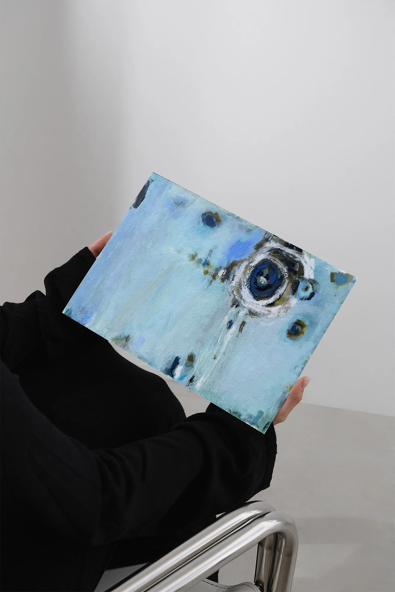 Person holding a pale blue abstract painting with a dark circular motif and layered textures, shown in a minimalist interior setting.