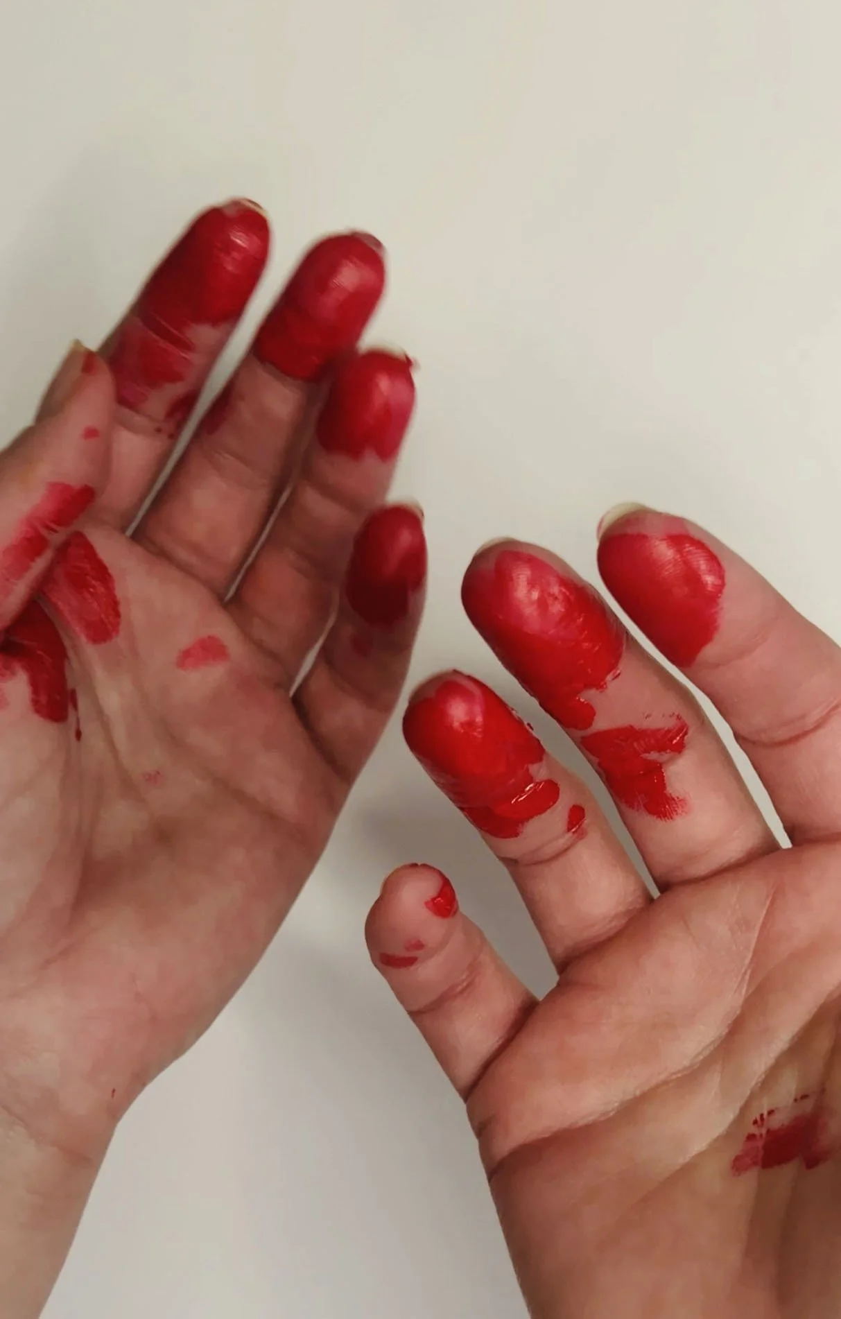Close-up of two hands covered in vivid red paint, fingers partially coated and stained, capturing a spontaneous, tactile creative process.