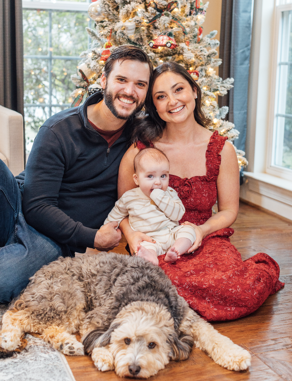 Couple sitting on the floor holding six month old baby with puppy in front of them. They are sitting in front of their Christmas tree.