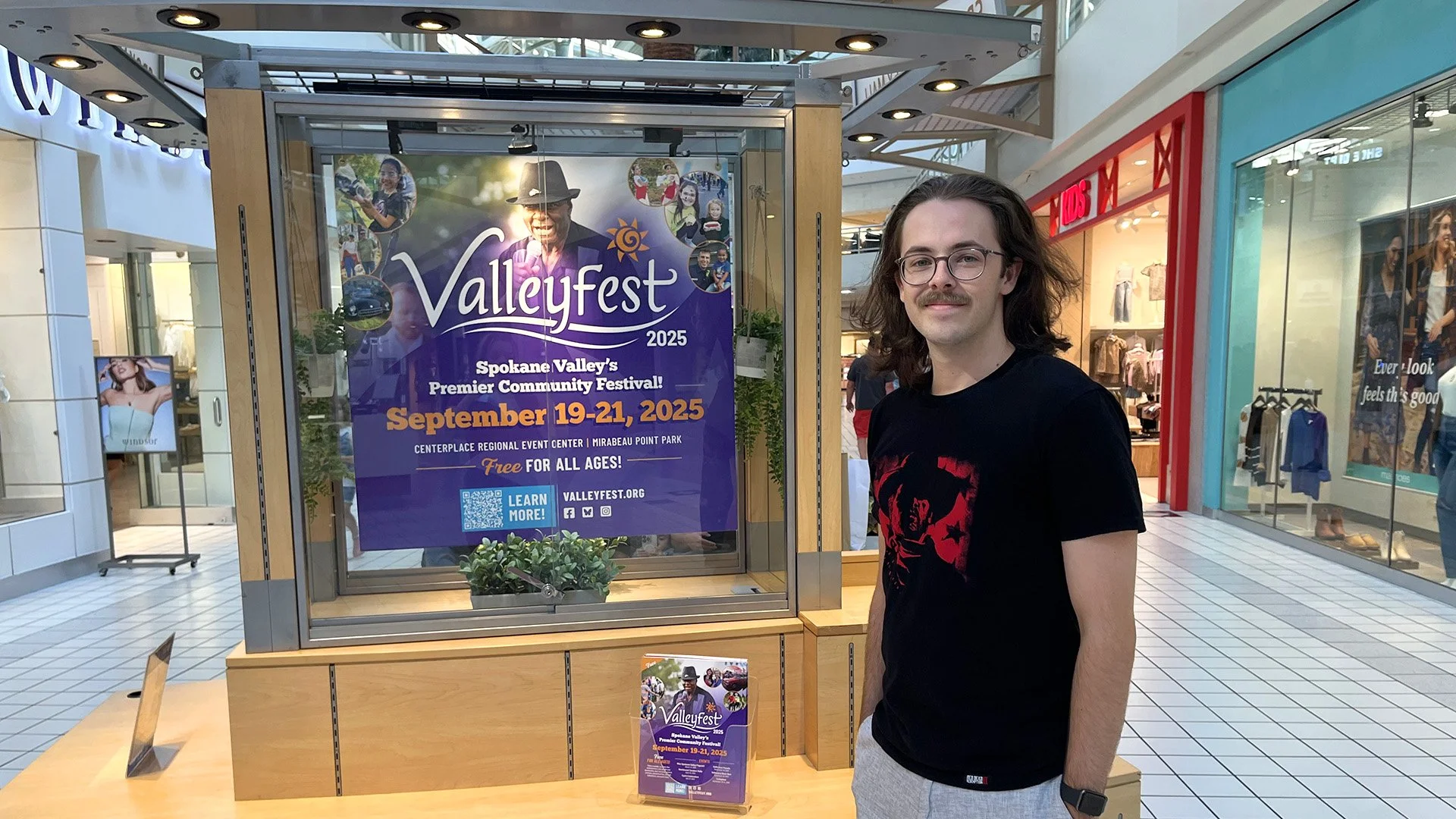 Photograph of Connor Woog posing with a Valleyfest display at the Spokane Valley Mall. This display consists of posters, flyers, etc.