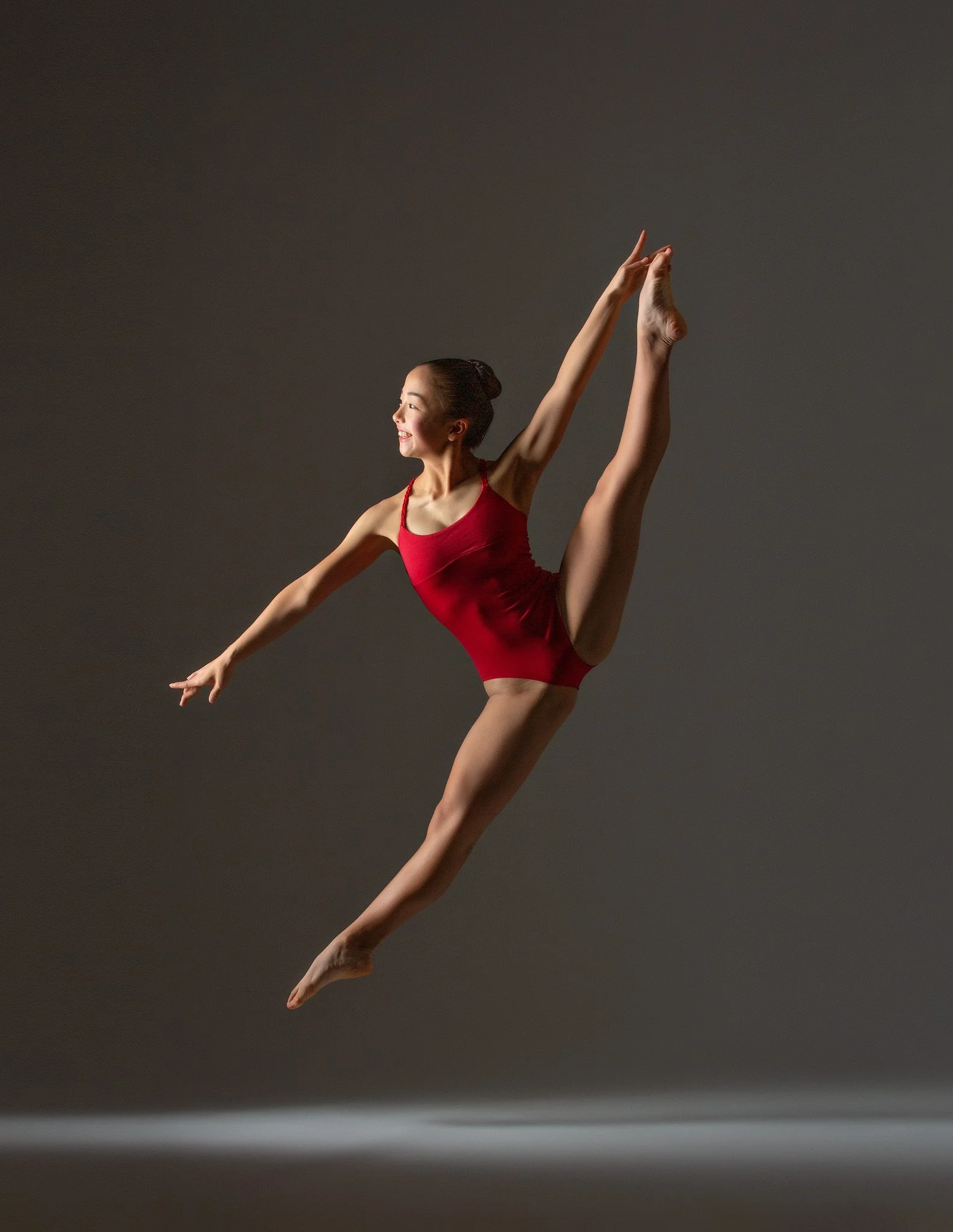 A female gymnast in a red leotard performing a leap against a gray background.
