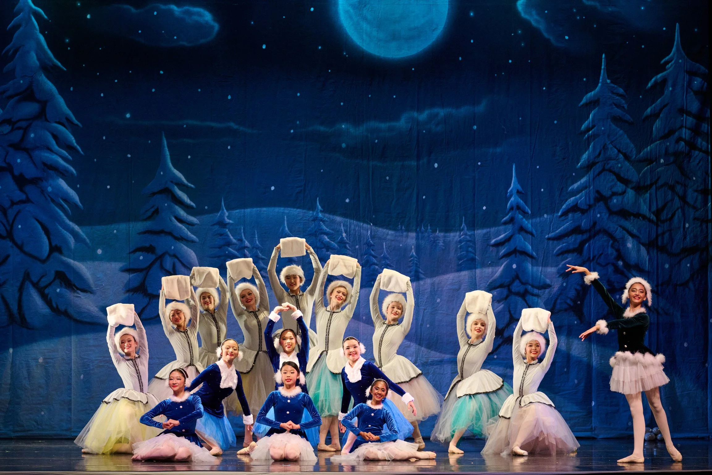 A group of ballet dancers in costumes performing on stage with a winter scene backdrop showing snow-covered trees and a moonlit night.