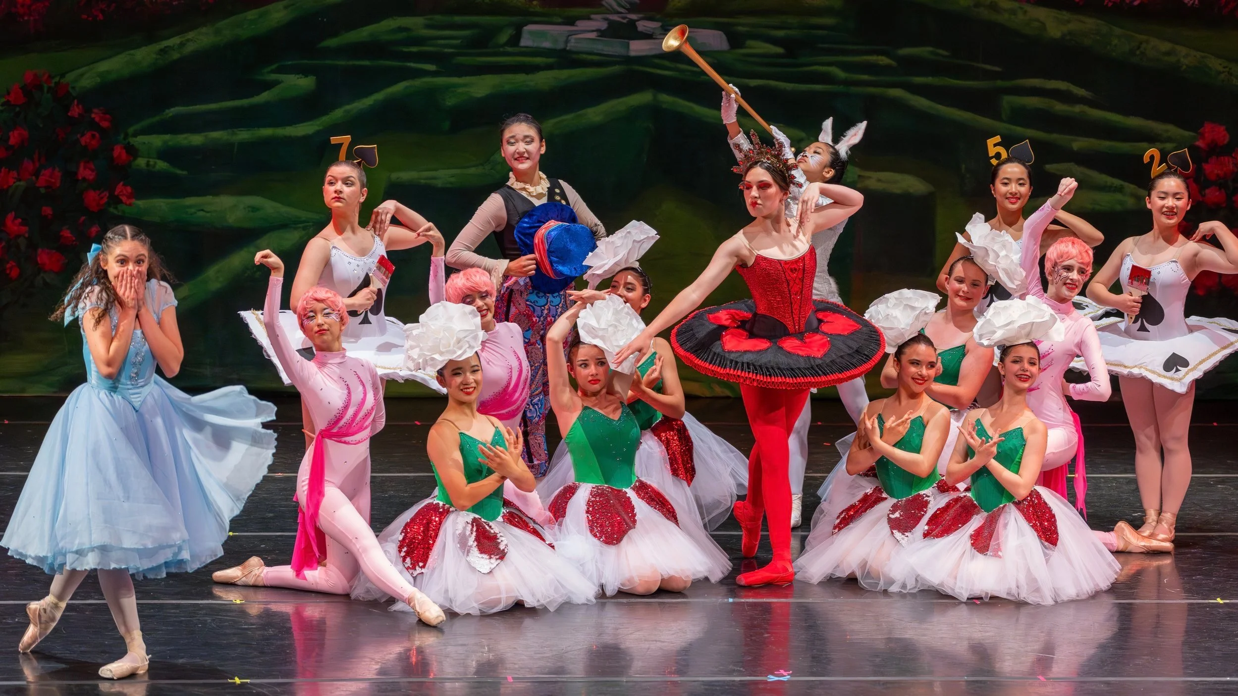 Ballet dancers on stage dressed in colorful costumes, some with pink and white, green and red, and others in blue, performing a scene with a painted forest background.