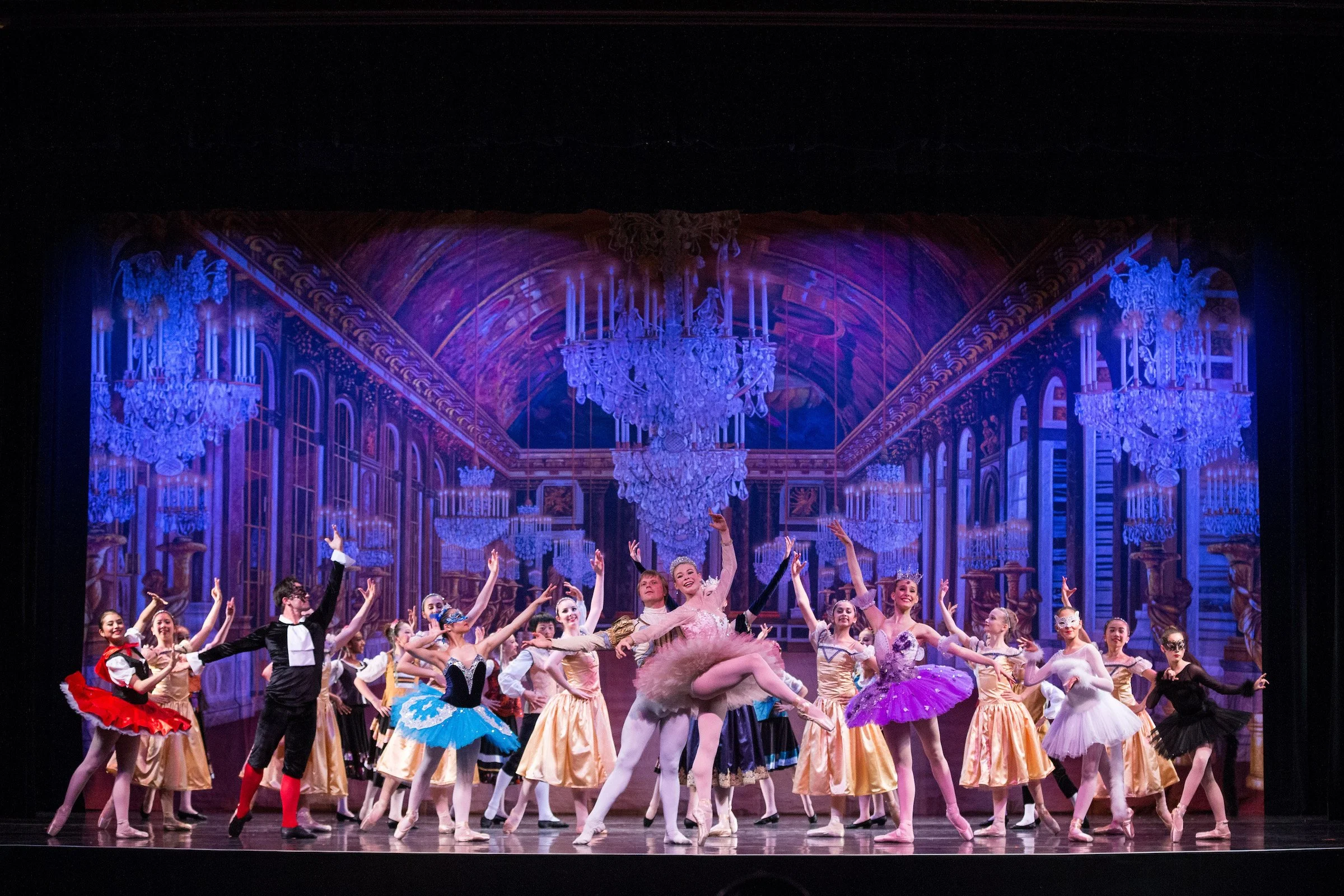 Ballet dancers performing on stage with a grand, elegant backdrop featuring chandeliers and ornate architecture.