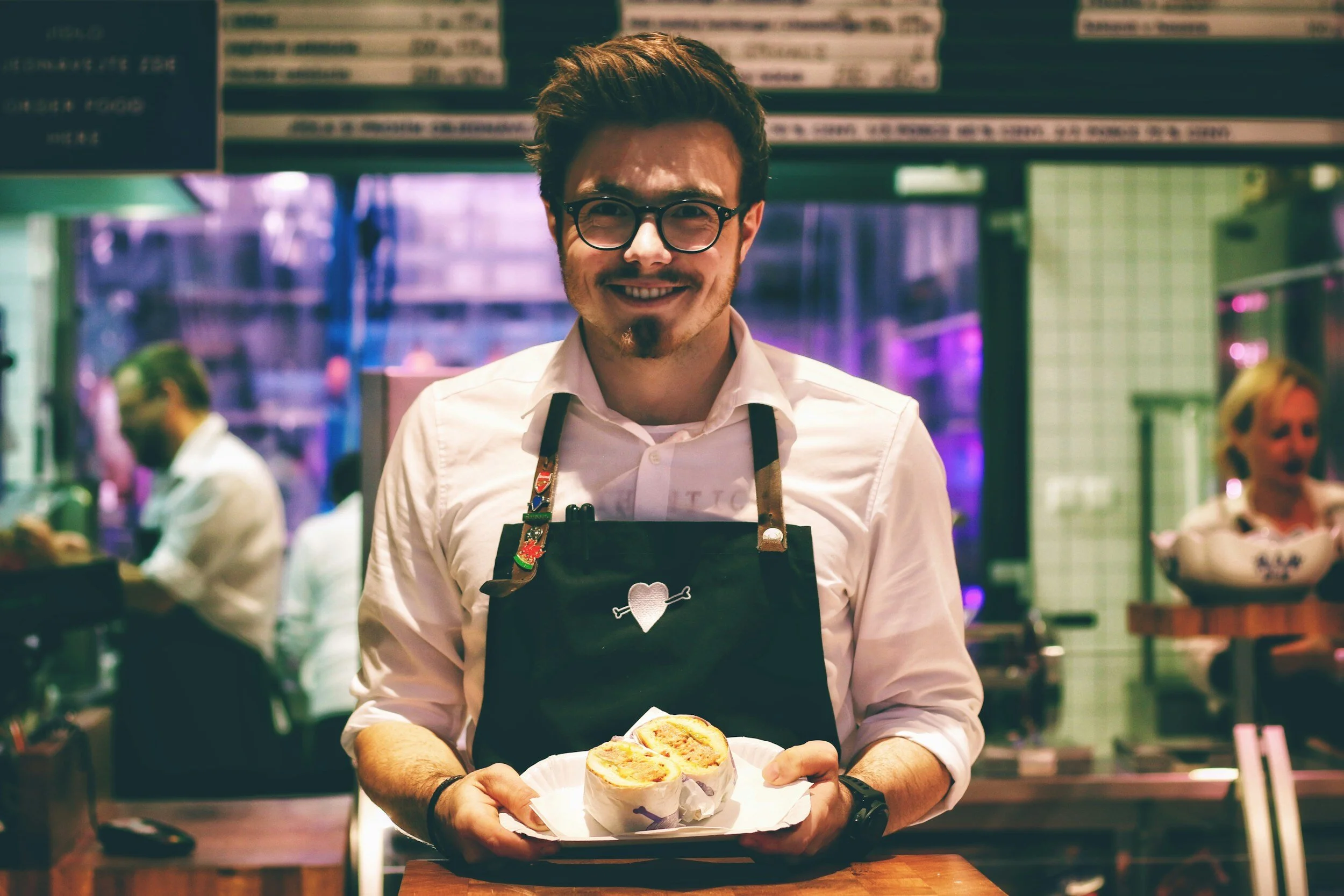 Smiling man in restaurant wearing apron holding sandwich.
