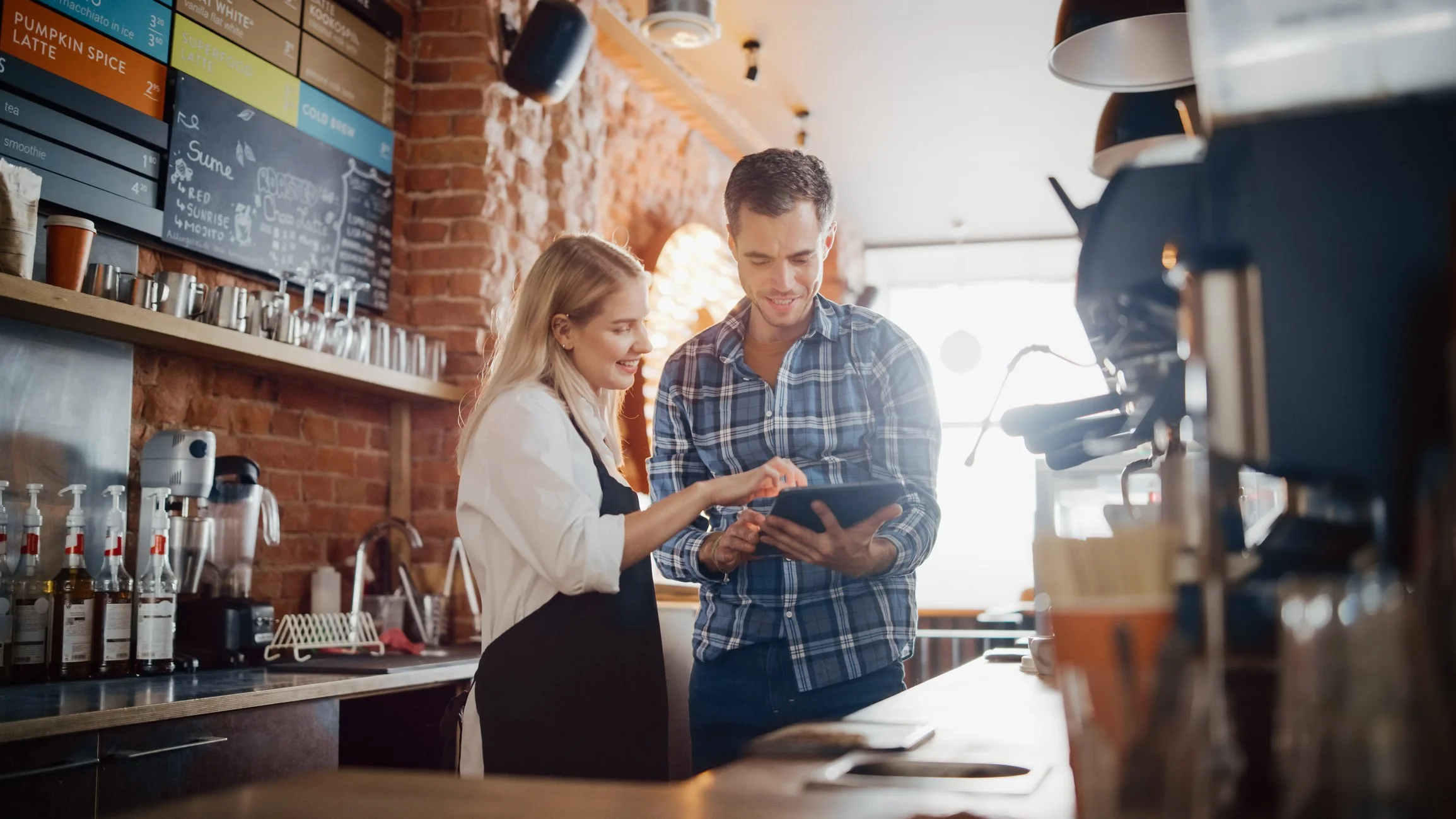 Two people in a cafe reviewing something on a tablet, surrounded by coffee shop equipment and menu, with a warm ambient light.