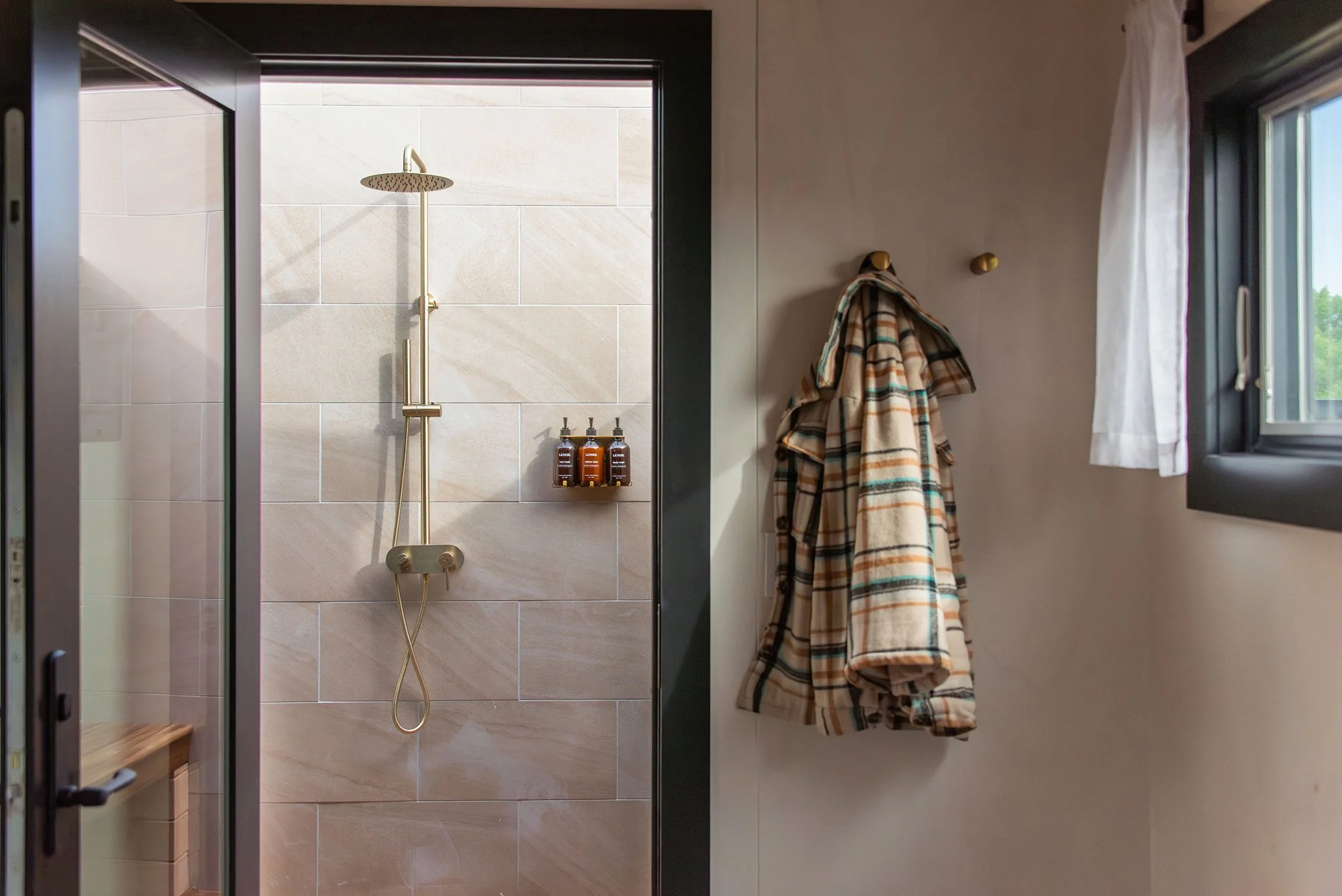 Deluxe Cabin - Bathroom with a private outdoor shower featuring a rain showerhead, bottles of soap and shampoo, and a hanging plaid coat near a window with a white curtain.