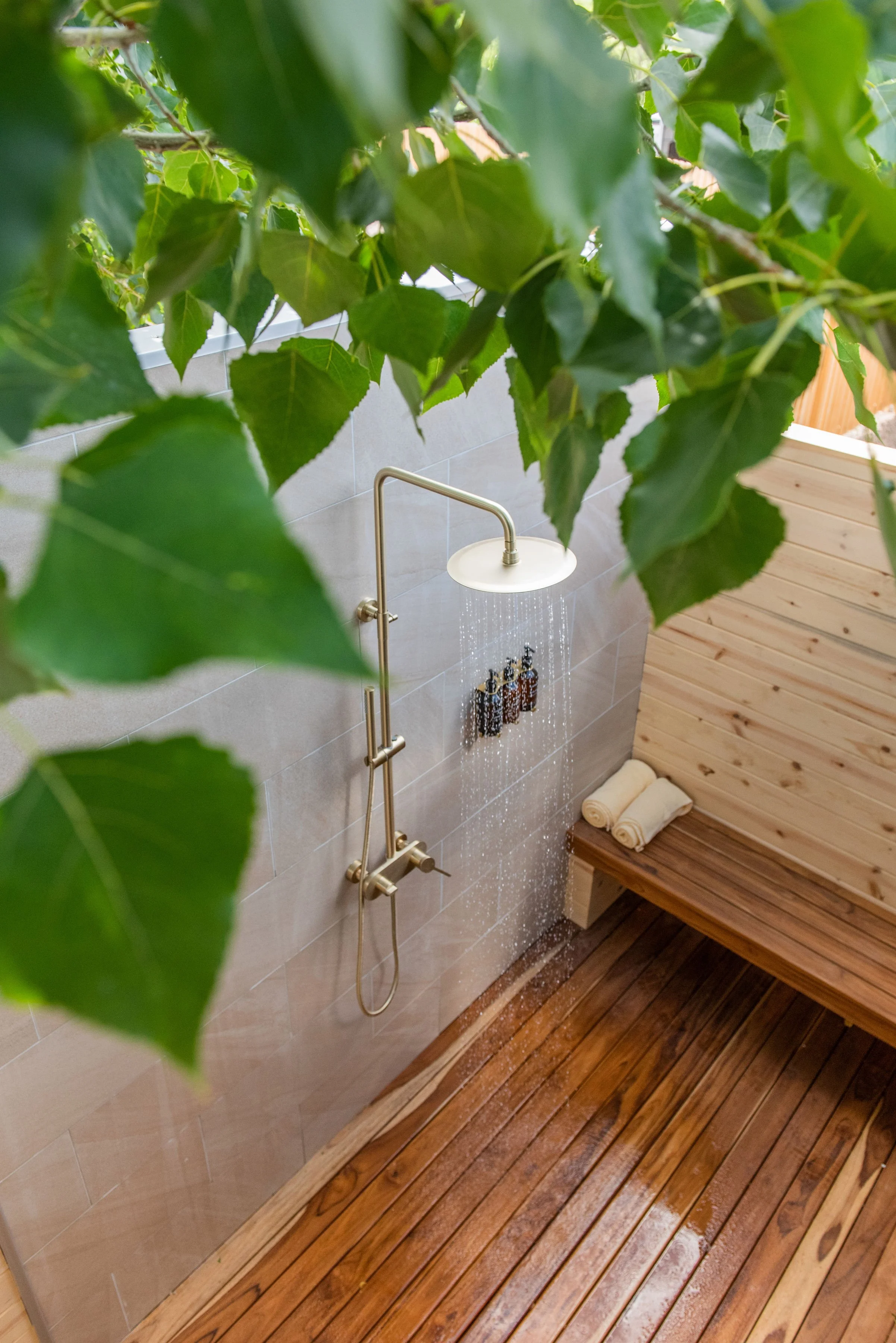 Private Outdoor Showers - An outdoor shower with a rainfall showerhead mounted on a grey tiled wall, surrounded by green foliage, and a wooden bench with rolled towels on a wooden deck.