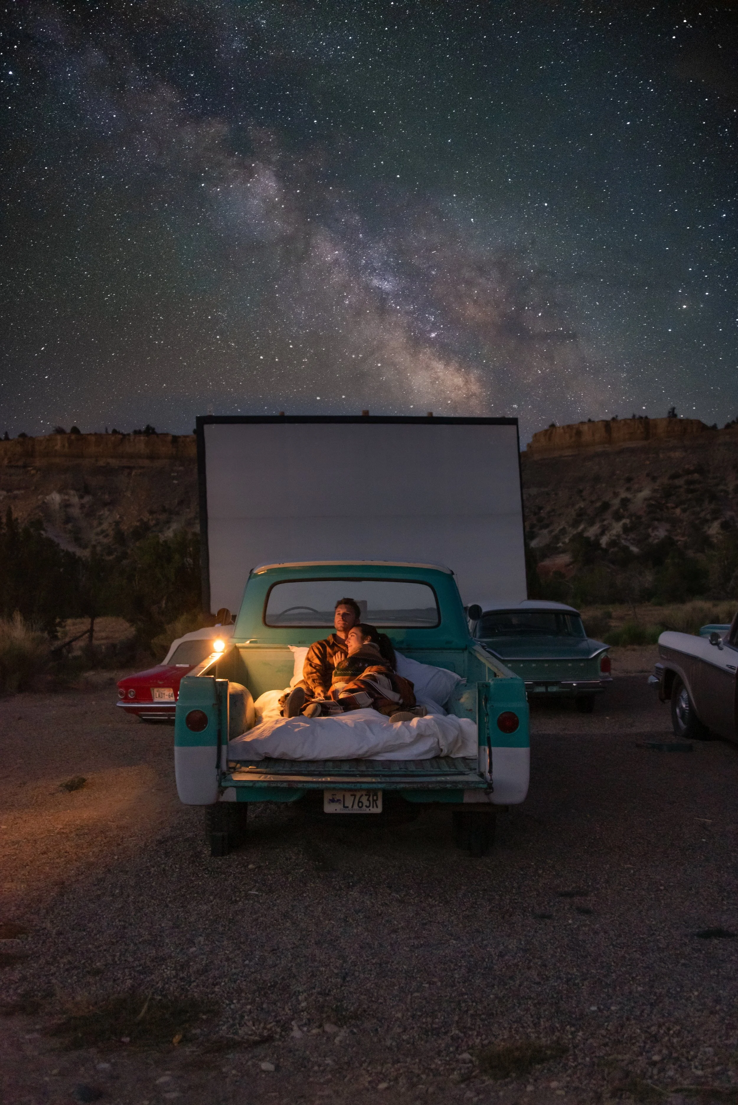 A couple sitting in the bed of a vintage turquoise pickup truck at night, under a starry sky with the Milky Way galaxy visible, surrounded by parked cars and rocky terrain.