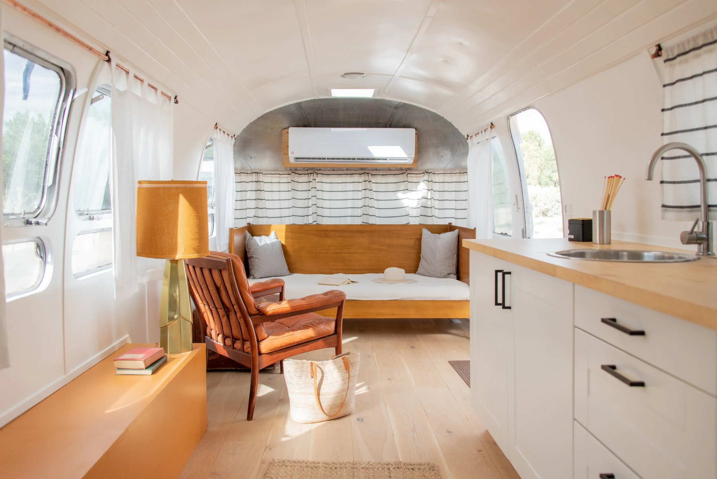 Vintage Airstream - Interior of an Airstream camper with a wooden bench, armchair, floor lamp, books, and a small kitchen area with a sink, drawers, and a countertop, all with white walls and windows with striped curtains.