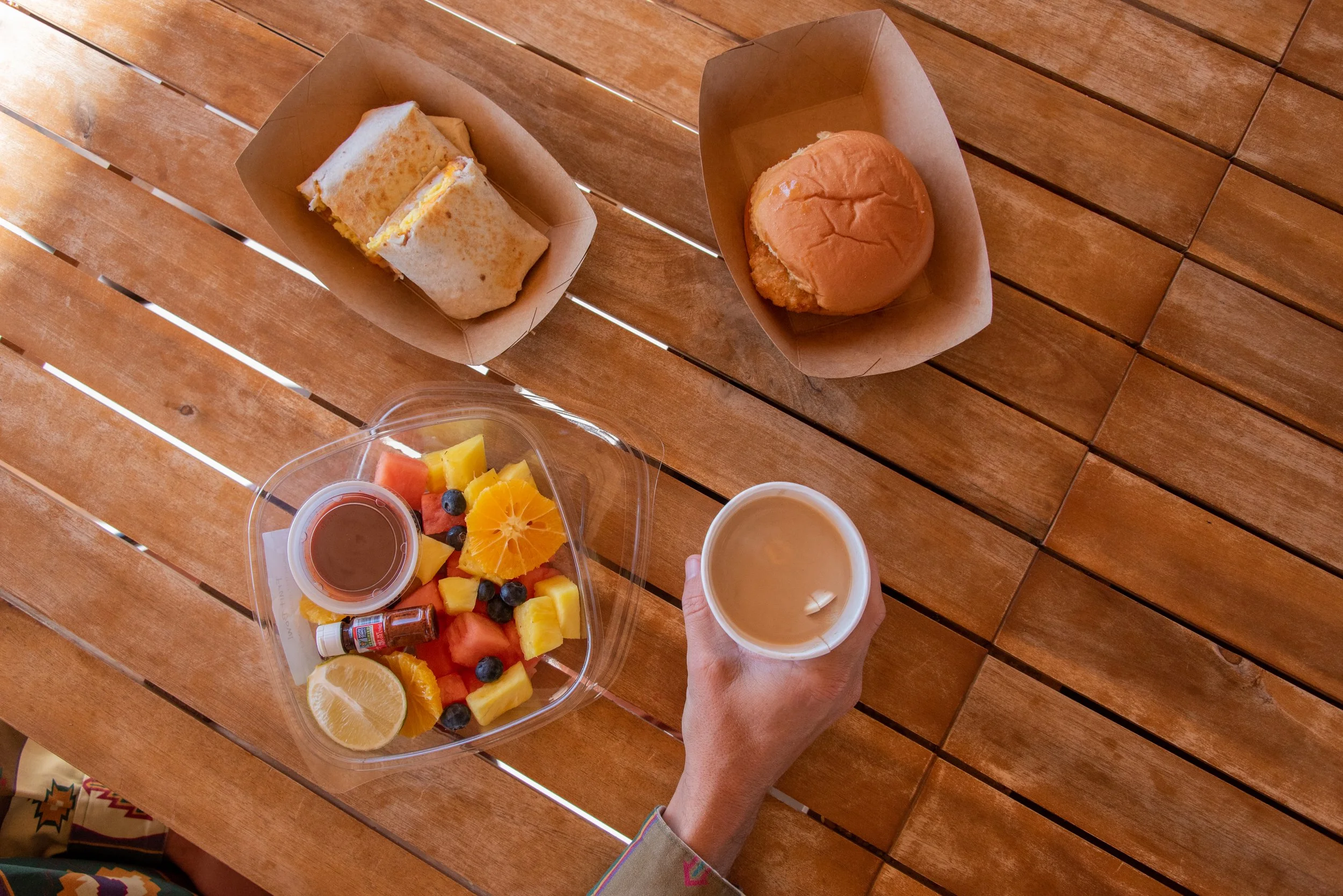 Overhead view of a table with a sandwich, a burger, a bowl of fruit salad, two cups of coffee, and a partially visible cup of orange juice on a wooden table.