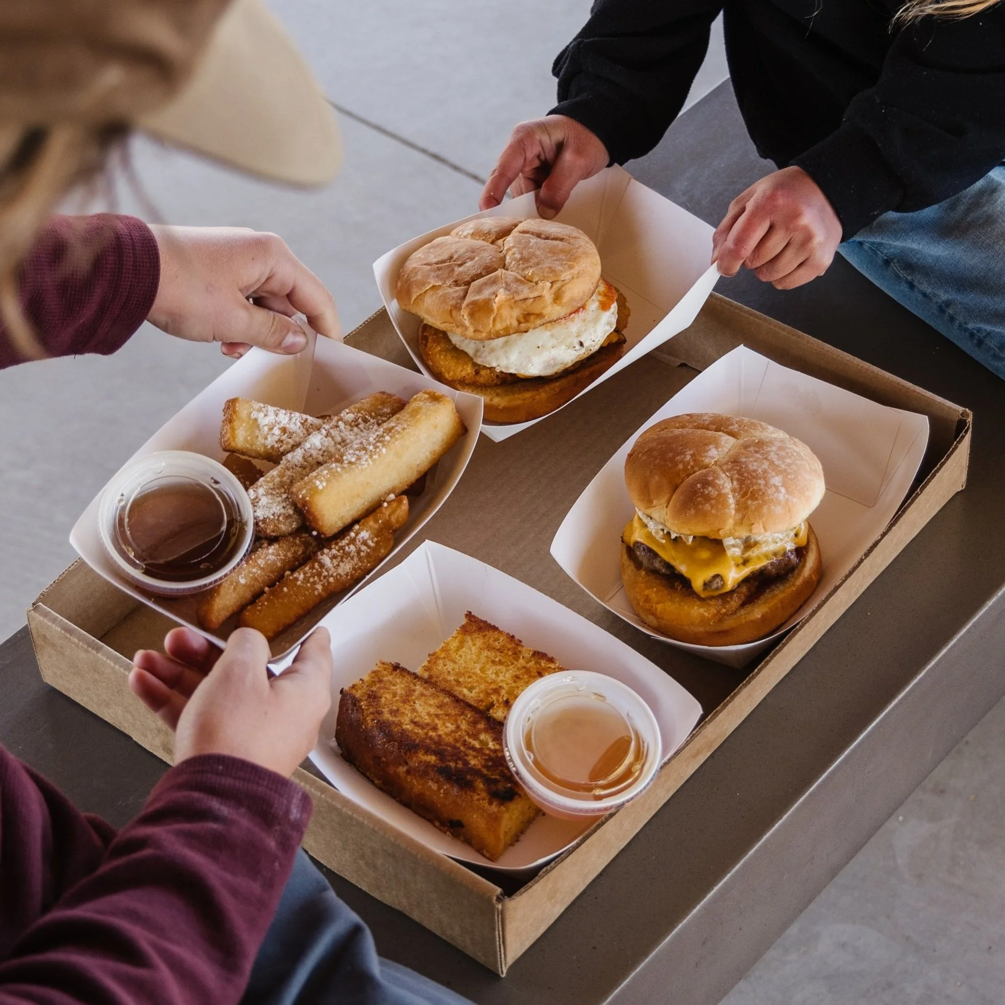 Two people holding a box of breakfast sandwiches and pastries, with small cups of syrup and dipping sauces on the table.