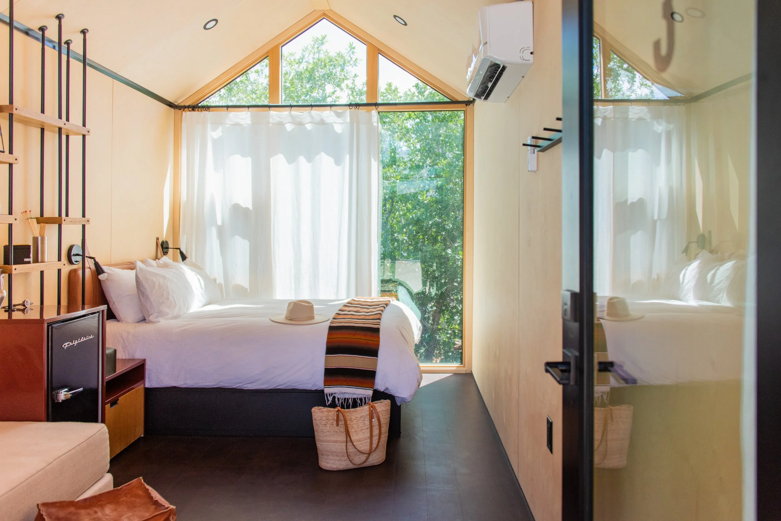 Tiny Cabin - A cozy bedroom with a bed against the left wall, topped with white pillows and a striped blanket. The room has large front-facing windows with white curtains, allowing natural light in. Air conditioner pictured. 