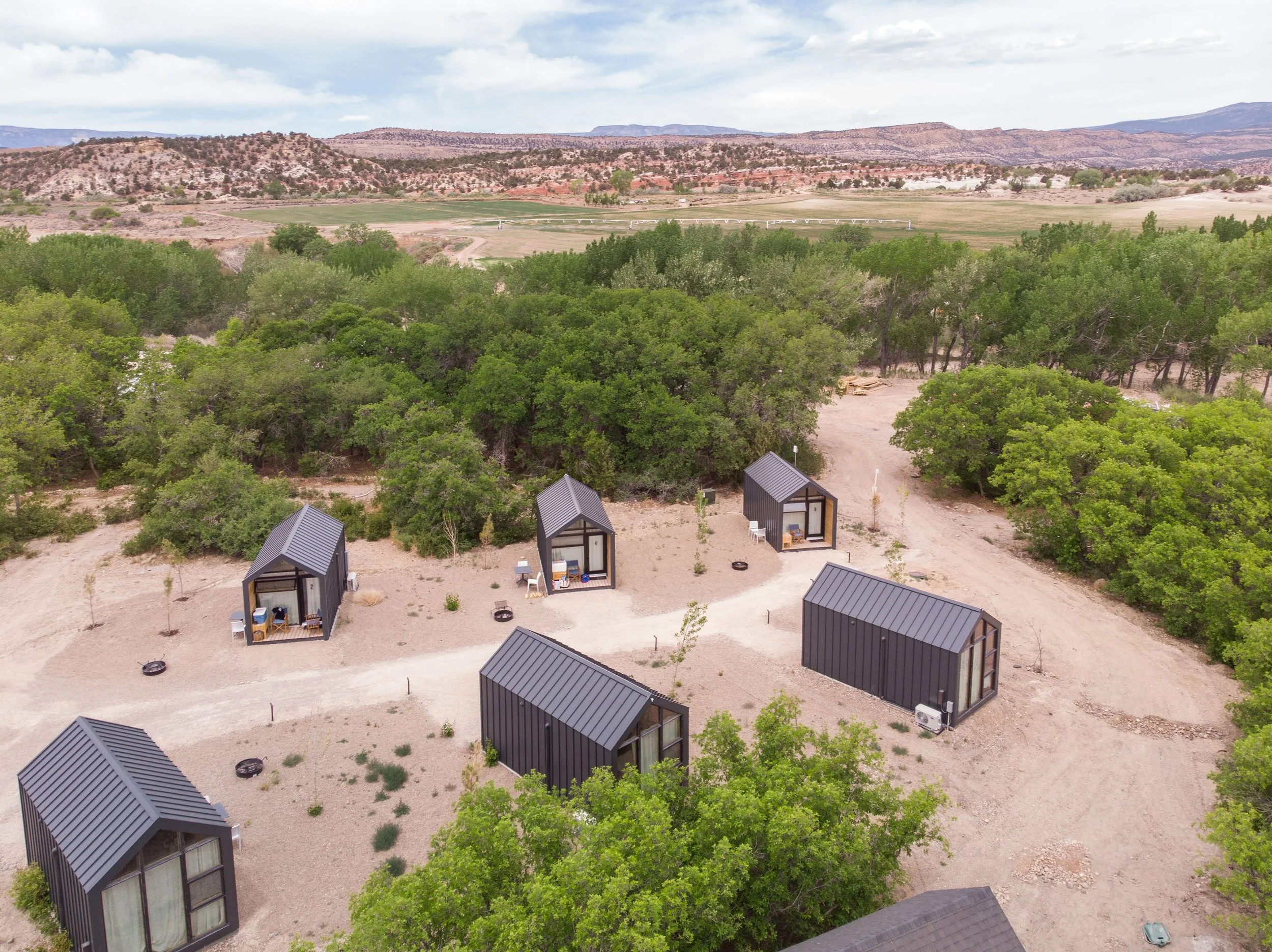 Aerial view of tiny cabins with metal roofs arranged in a nature setting with trees and open land and distant hills in the background.