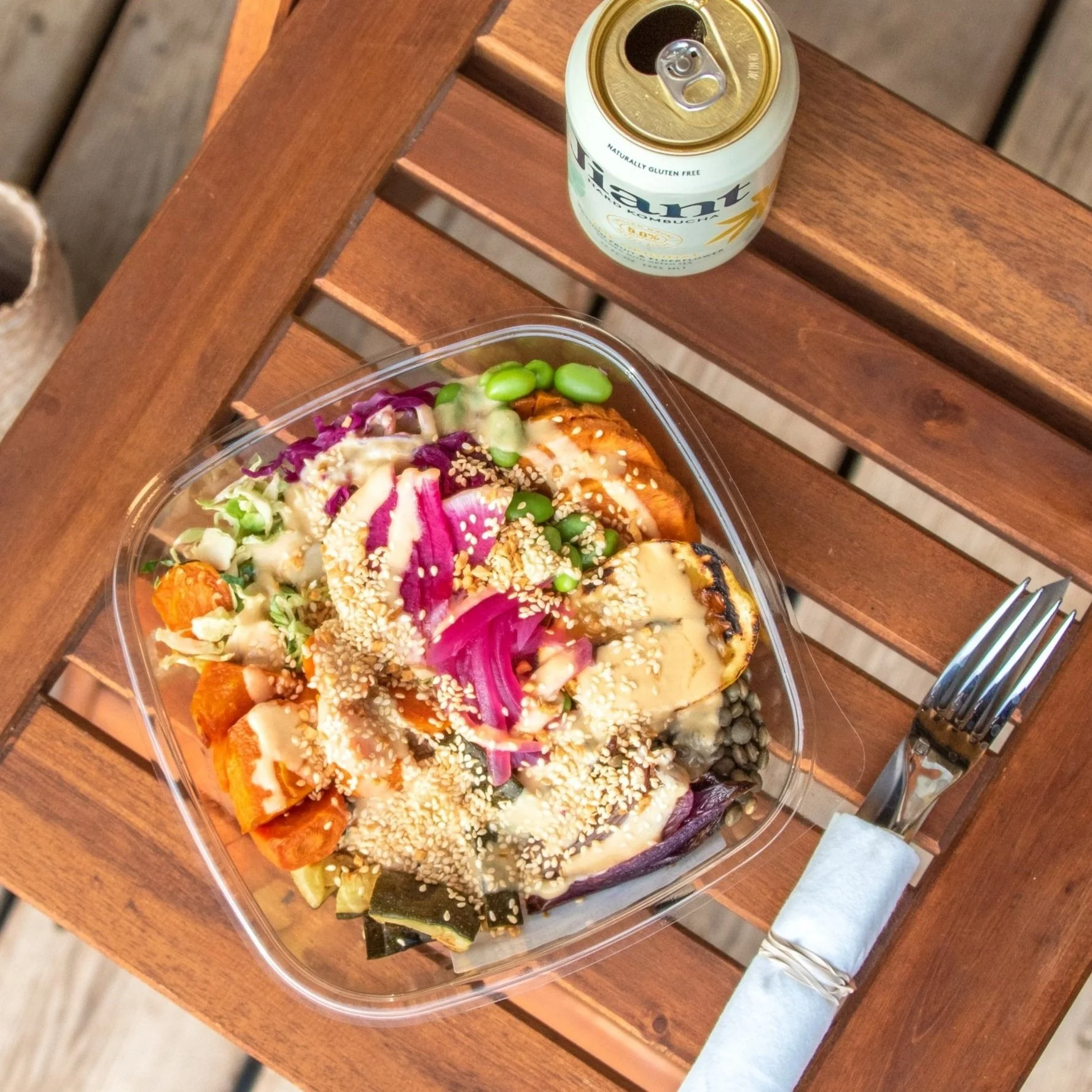 A salad with grilled chicken, edamame, purple cabbage, carrots, sesame seeds, and avocado in a clear container on a wooden table with a fork wrapped in a napkin beside it, and a can of LaCroix sparkling water in the background.
