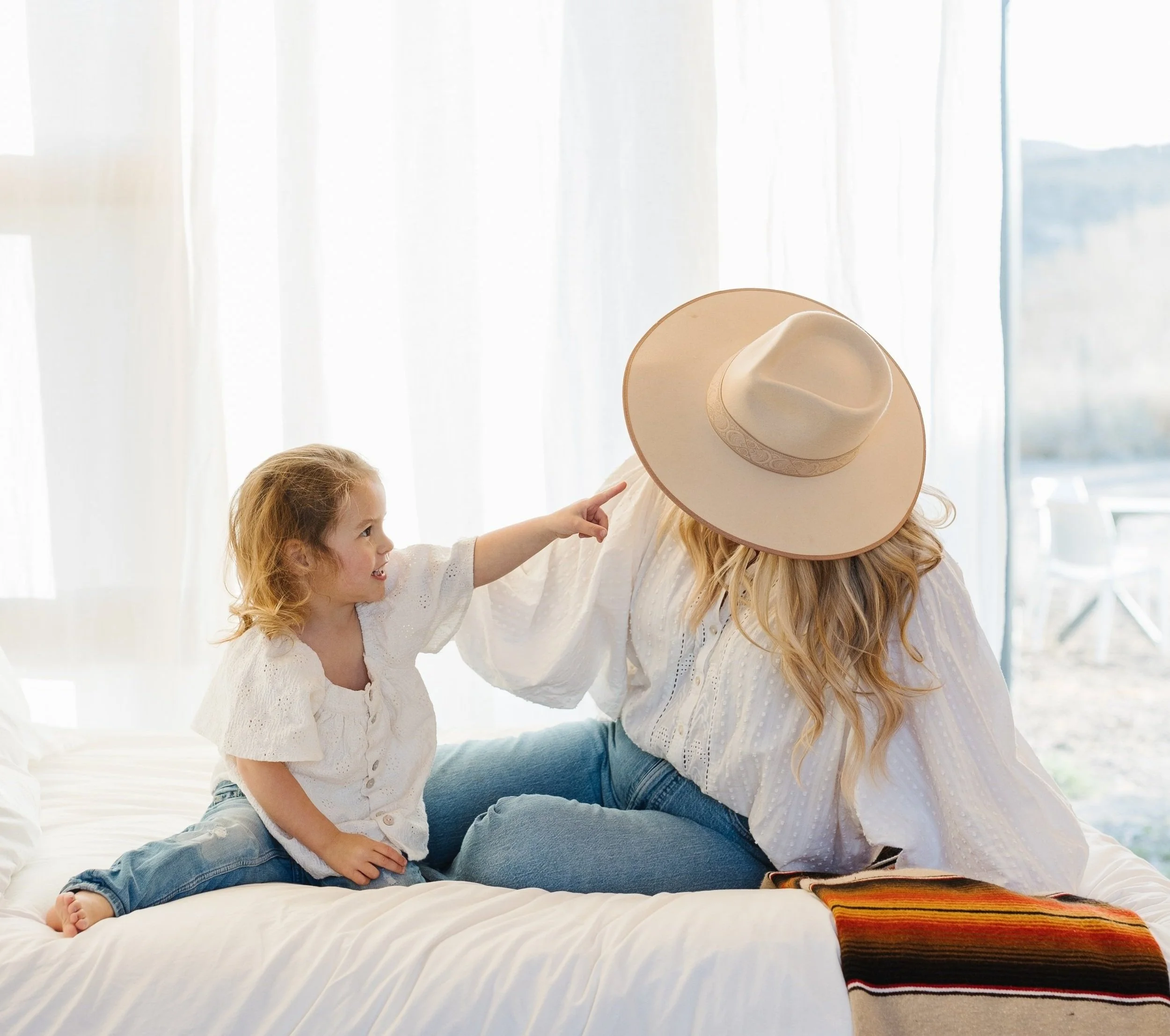 A woman with long blonde hair wearing a wide-brimmed hat sitting on a bed with a young girl with curly blonde hair, both smiling and reaching toward the woman's hat. They are in a bright room with light-colored curtains. Ofland family friendly