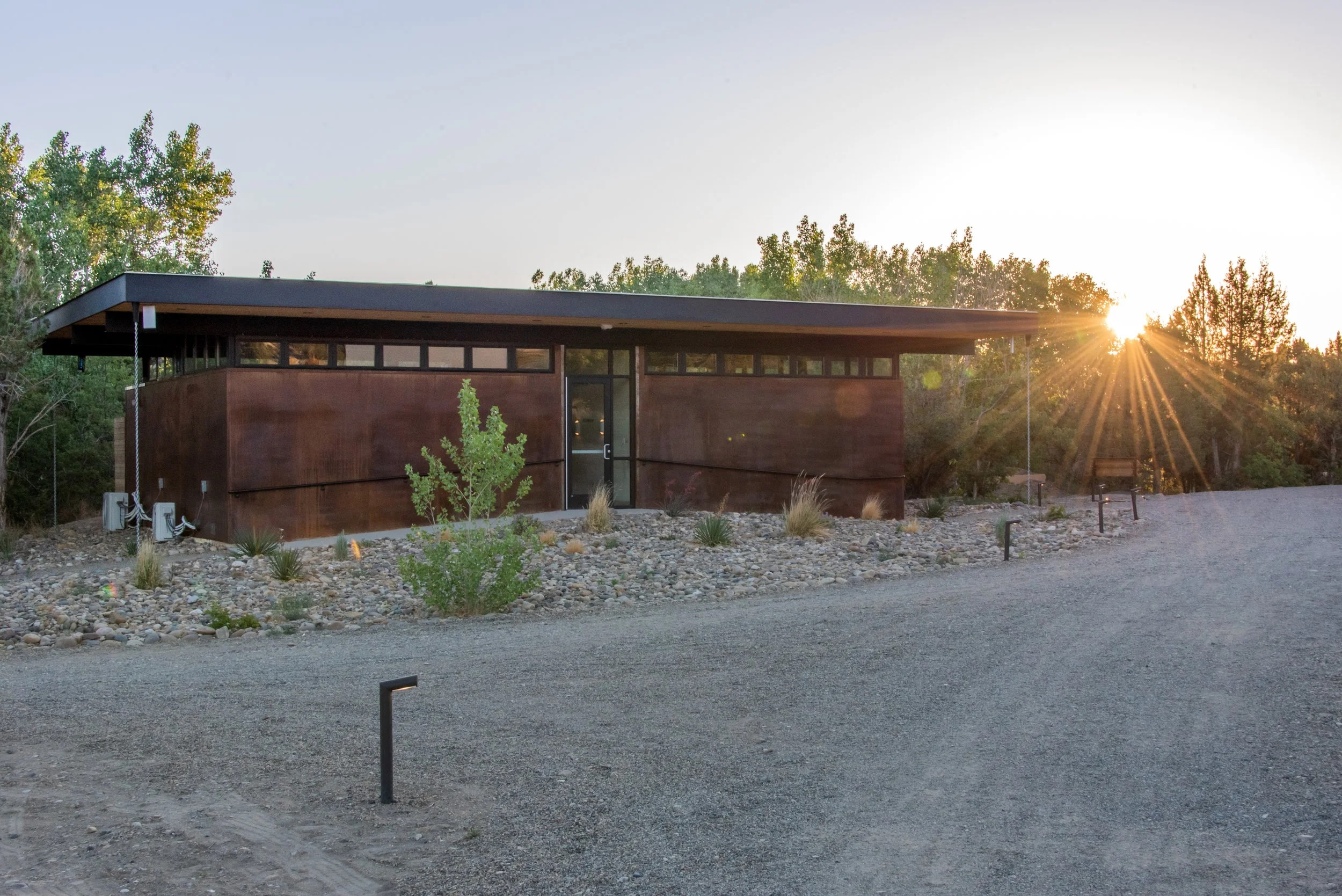 Spa-Quality Bathhouses - A modern, single-story building made of rust-colored metal with a flat roof, located in a natural setting with gravel and desert plants, as the sun sets behind trees in the background.