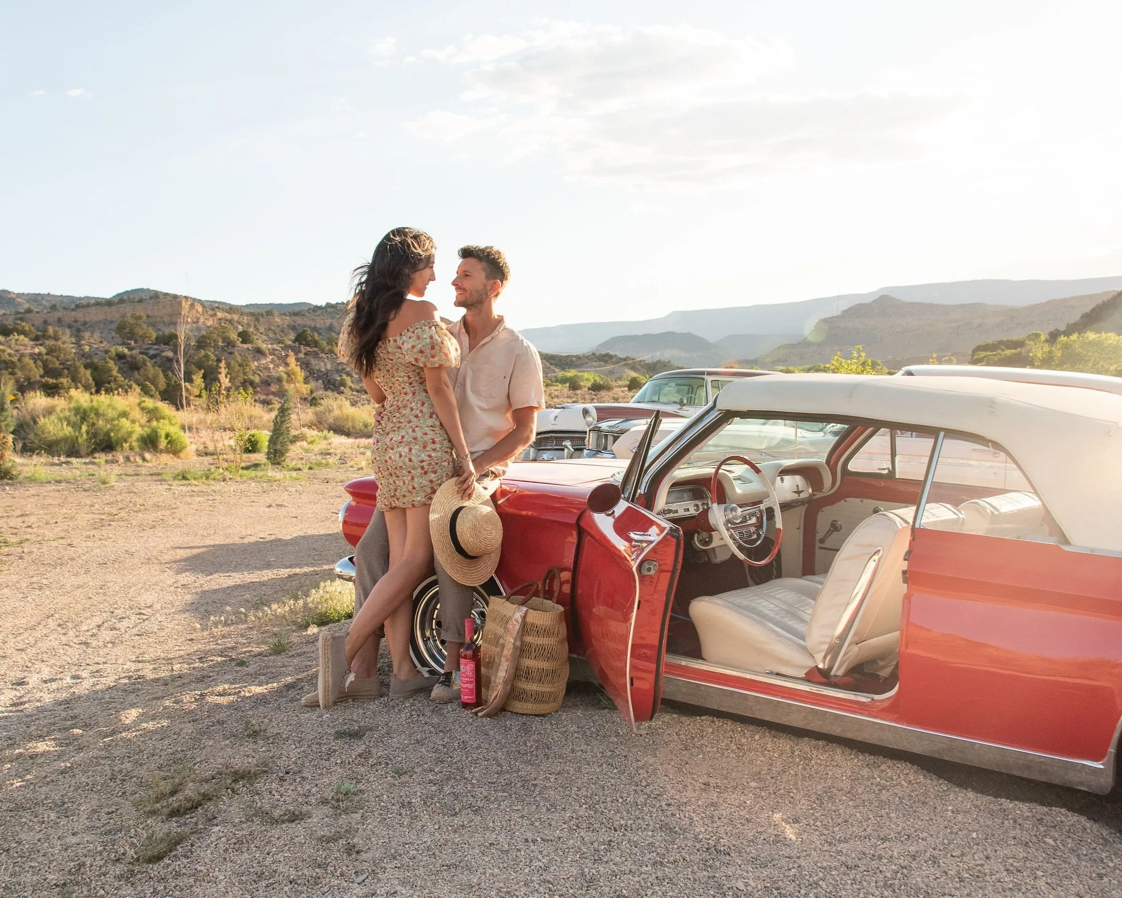 A young couple stands beside a vintage red car in a rural landscape, holding hands and looking into each other's eyes as the sun sets behind mountains. Ofland couples friendly