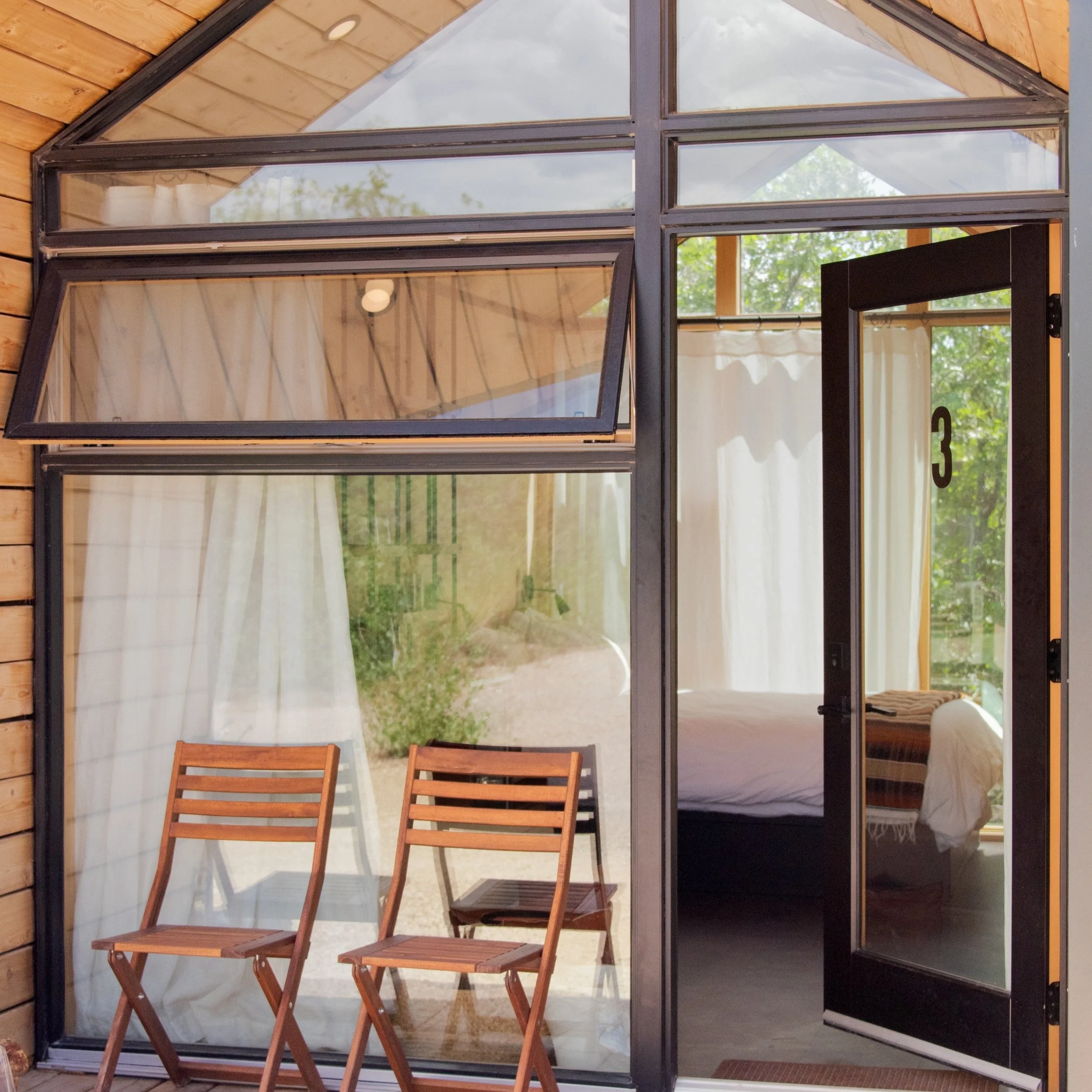 Tiny Cabin - View of a modern glass house with an open door. Two wooden chairs and a small wooden table outside near large glass windows and curtains inside. Green trees and garden seen through the windows.