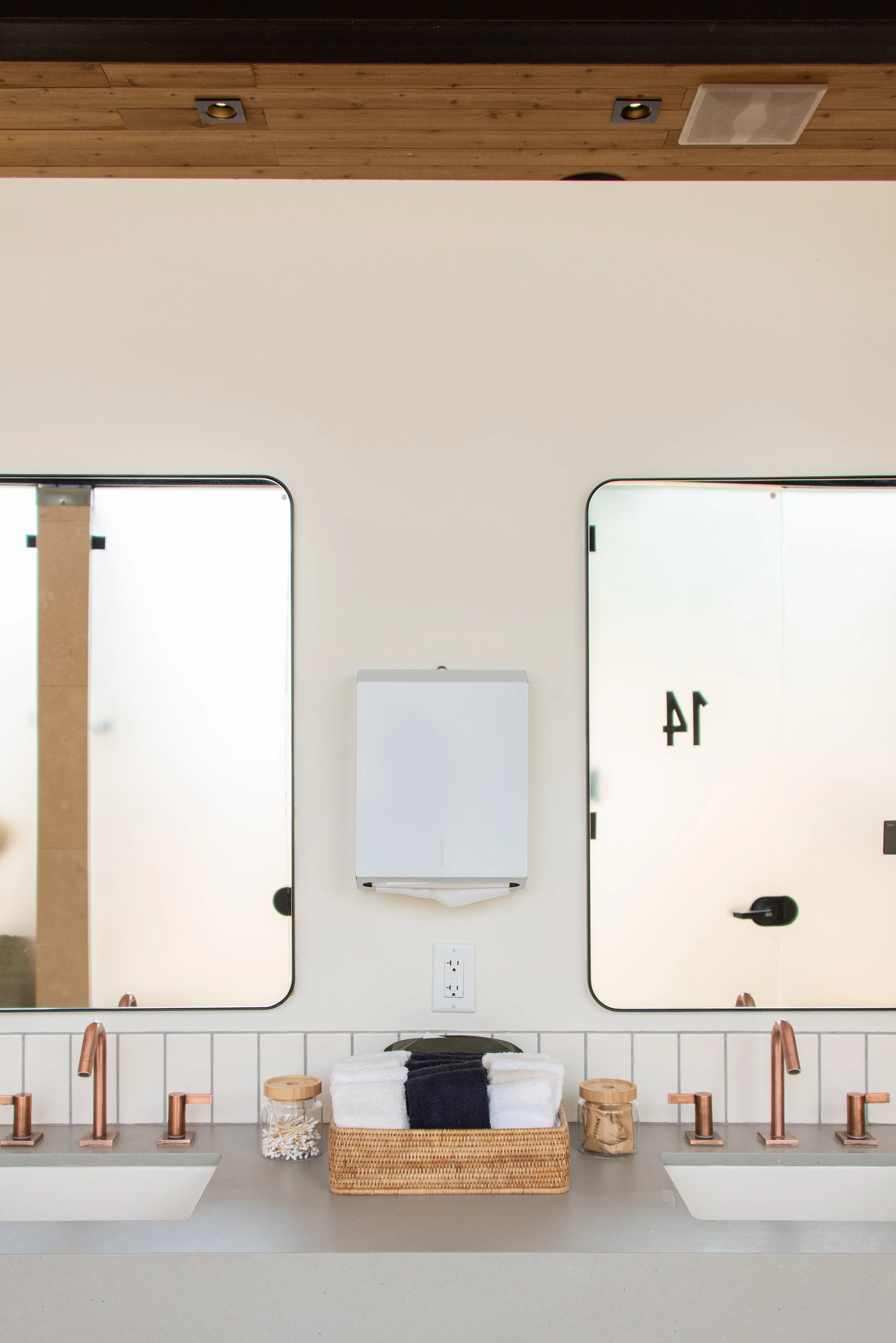 Spa-Quality Bathhouses - Bathroom with two mirrors, two sinks with copper faucets, a hand towel in a basket, and some small jars with Q-Tips, washcloths, and cotton rounds on a beige countertop.