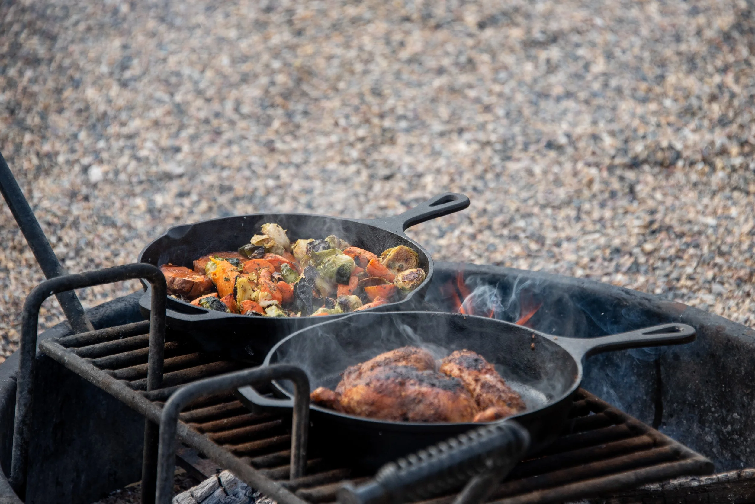 DIY Meal Kits - Two cast iron skillets cooking over an outdoor grill, one with roasted vegetables and the other with a grilled piece of meat, with visible smoke rising.
