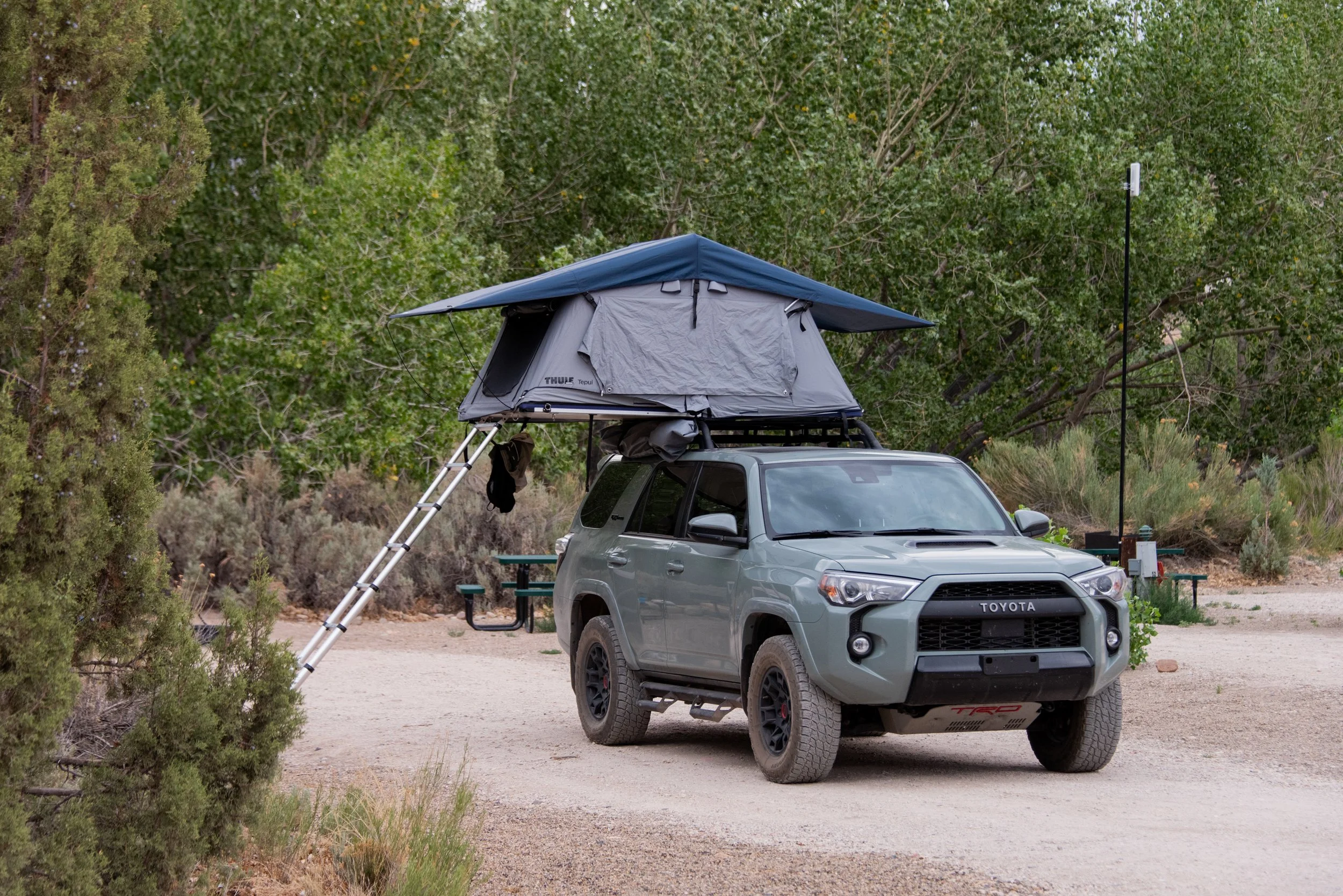 Campsite - A gray Toyota SUV equipped with a rooftop tent and an extended awning, parked on a dirt path in a natural, wooded area.
