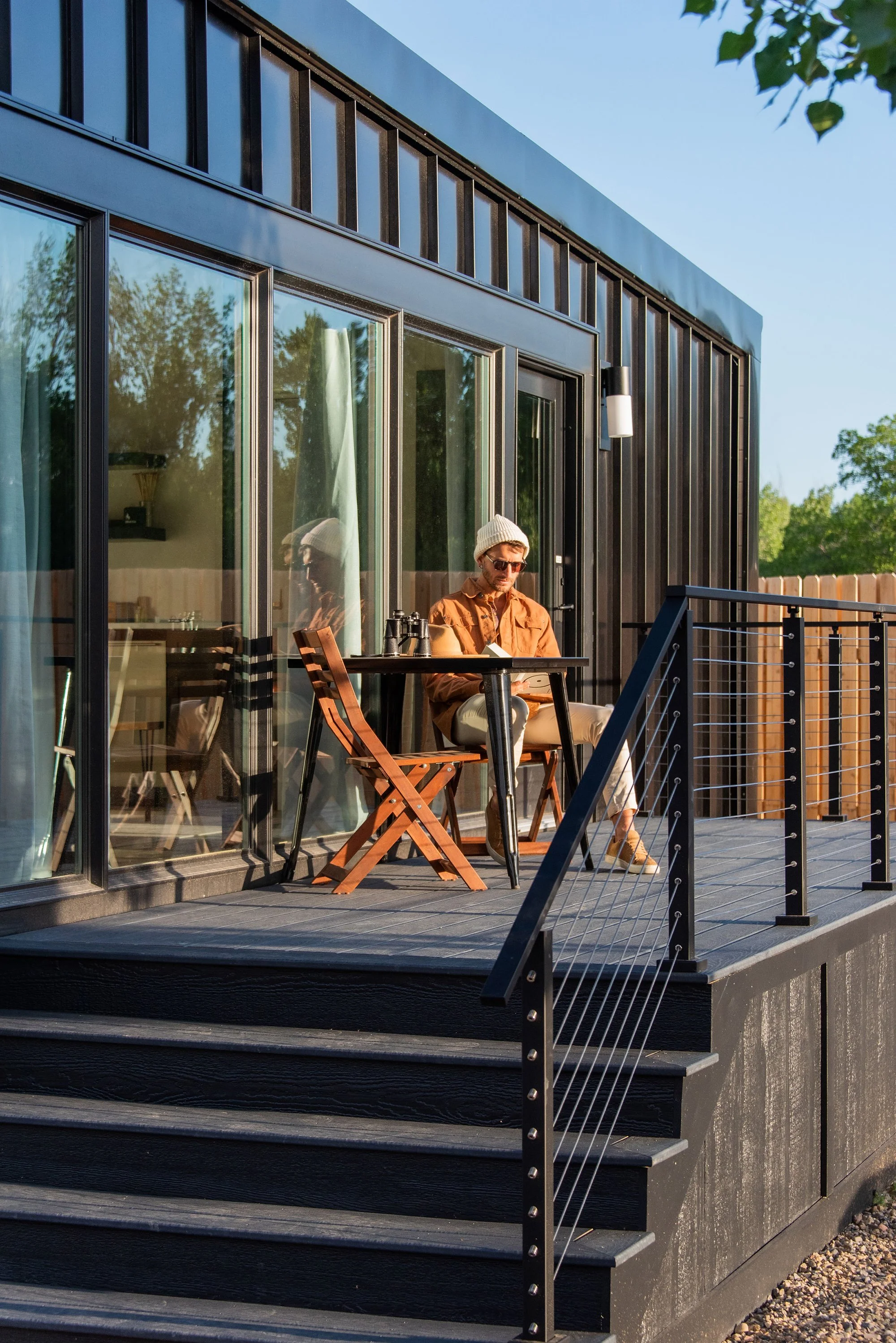 Deluxe Cabin - A man sitting on a patio balcony with a table and chairs, reading a book, wearing sunglasses, a beanie, and a brown jacket, next to a modern black house with large glass sliding doors and a railing, during sunset.