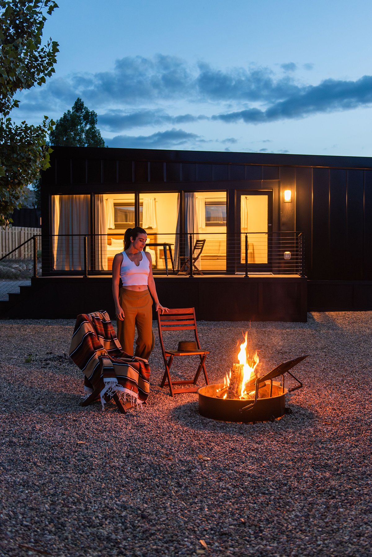 Deluxe Cabin - A young woman standing next to a campfire at dusk outside a modern cabin with large windows and a balcony.