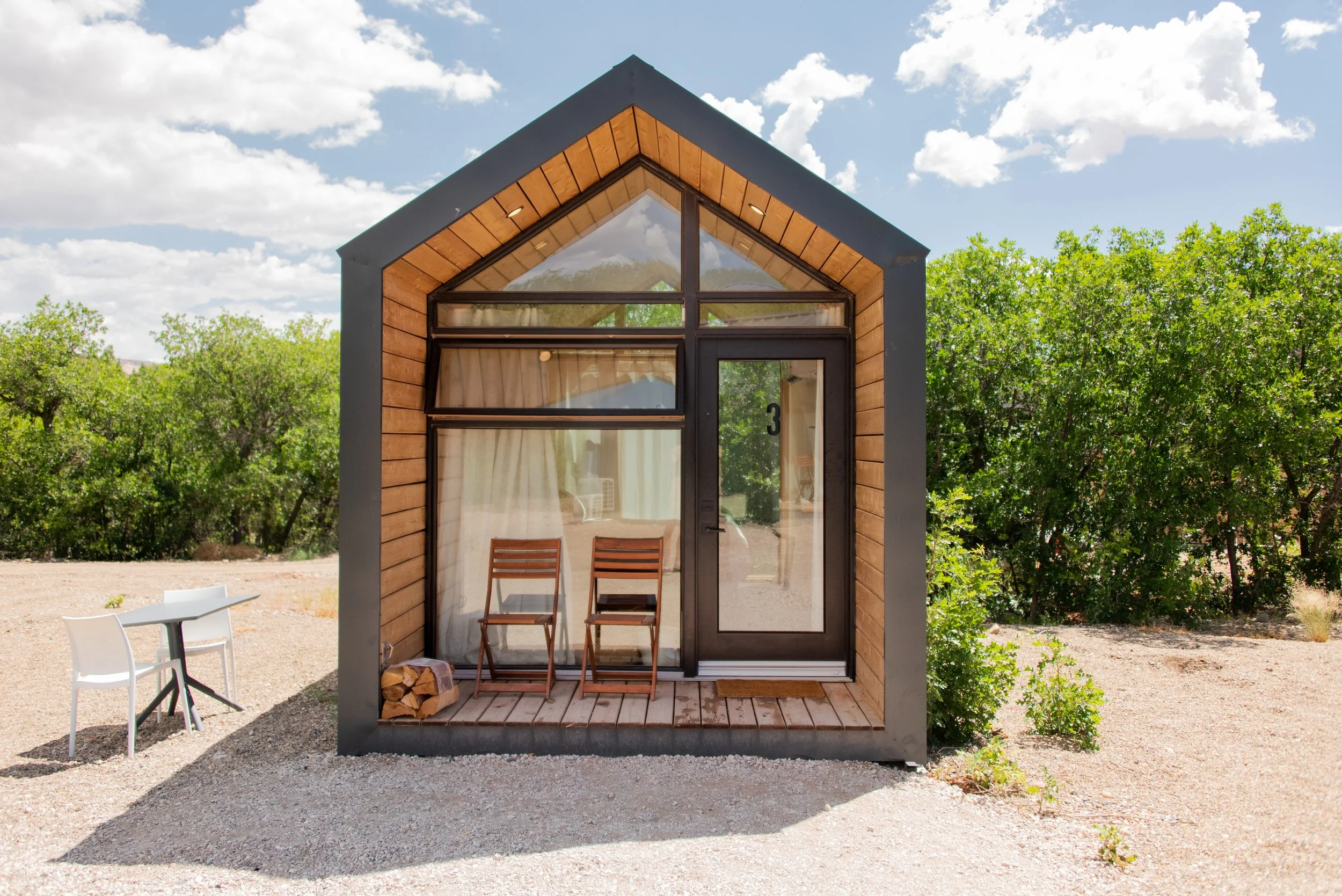 Tiny Cabin - Modern tiny cabin with wood and glass exterior, two chairs on porch, surrounded by greenery and trees, under partly cloudy sky. Sleeps 2 people. 