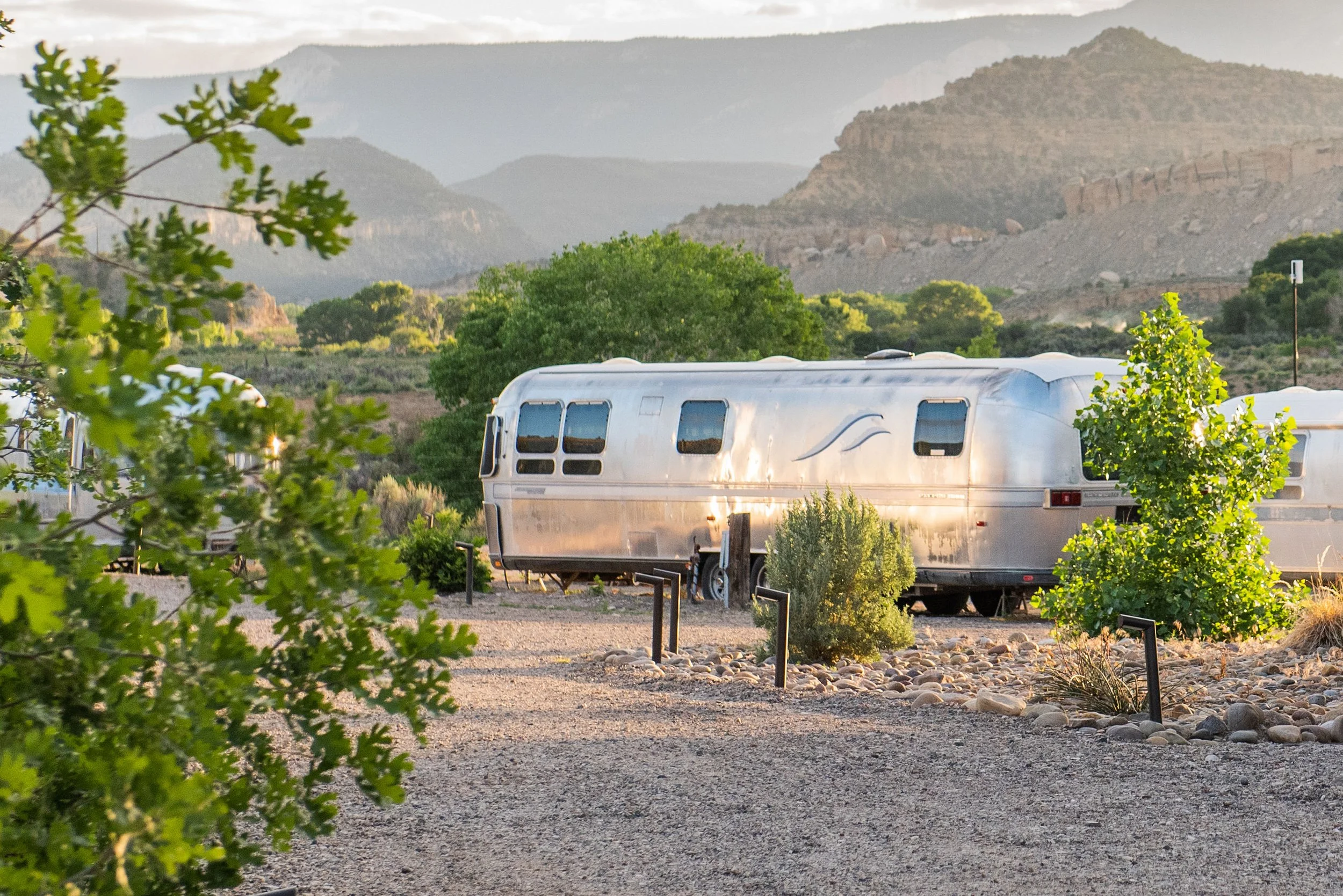 Vintage Airstream - A shiny vintage Airstream camper trailer parked in a desert landscape with mountains in the background and green bushes nearby during sunset.