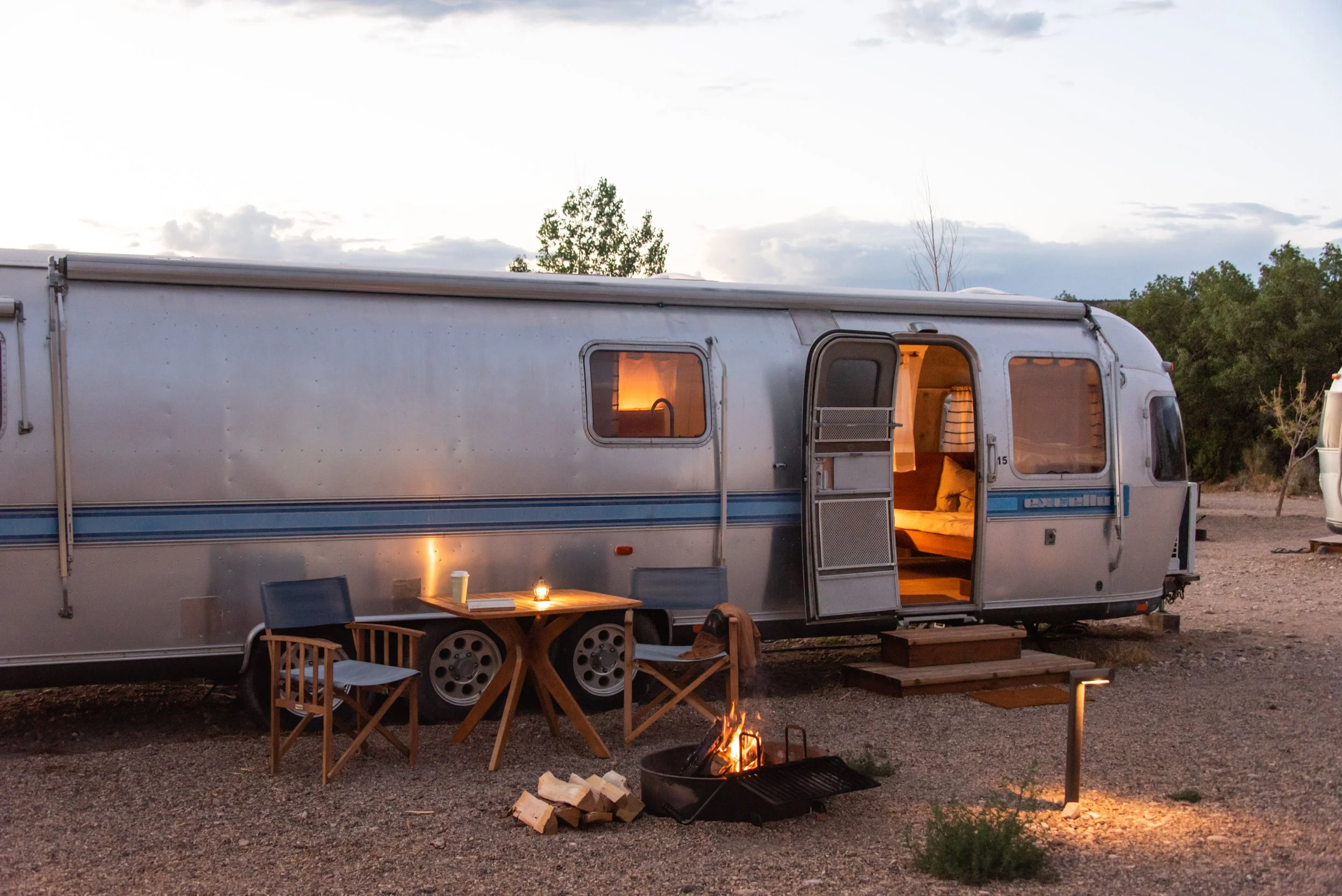 A vintage trailer parked outdoors during dusk, with warm interior lighting visible through the windows. In front, there are two wooden chairs, a small round table with a candle and coffee cup, and a campfire with stacked logs nearby. The scene depicts a cozy camping setup.