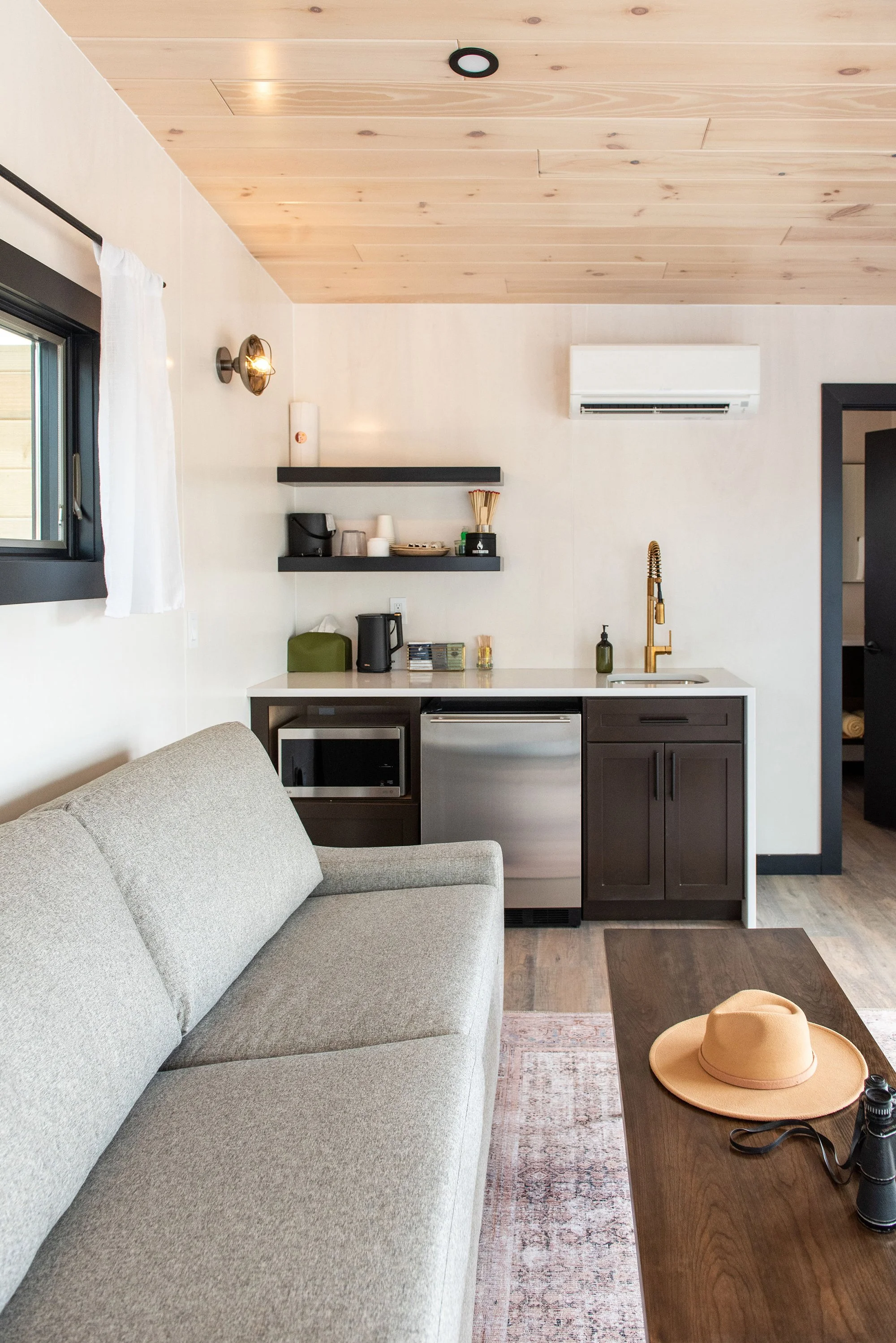 Deluxe Cabin - Interior of a cozy living space with a light beige sofa, a window with white curtains, a small kitchenette with a microwave, mini-fridge, and open shelves, and a wooden ceiling.