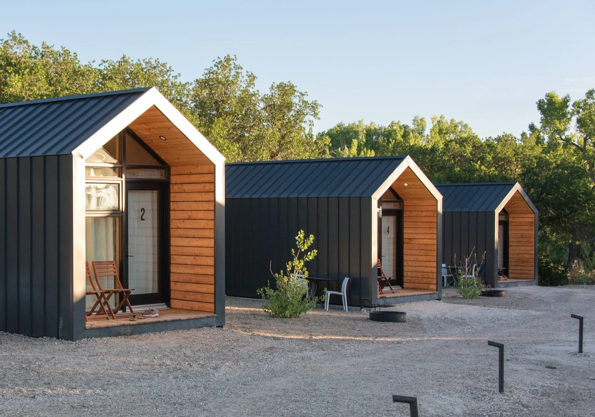 Tiny Cabins - Three modern tiny houses with black exteriors and wooden front accents, numbered 2, 4, and 6, situated outdoors on a gravel lot with trees in the background during sunrise.
