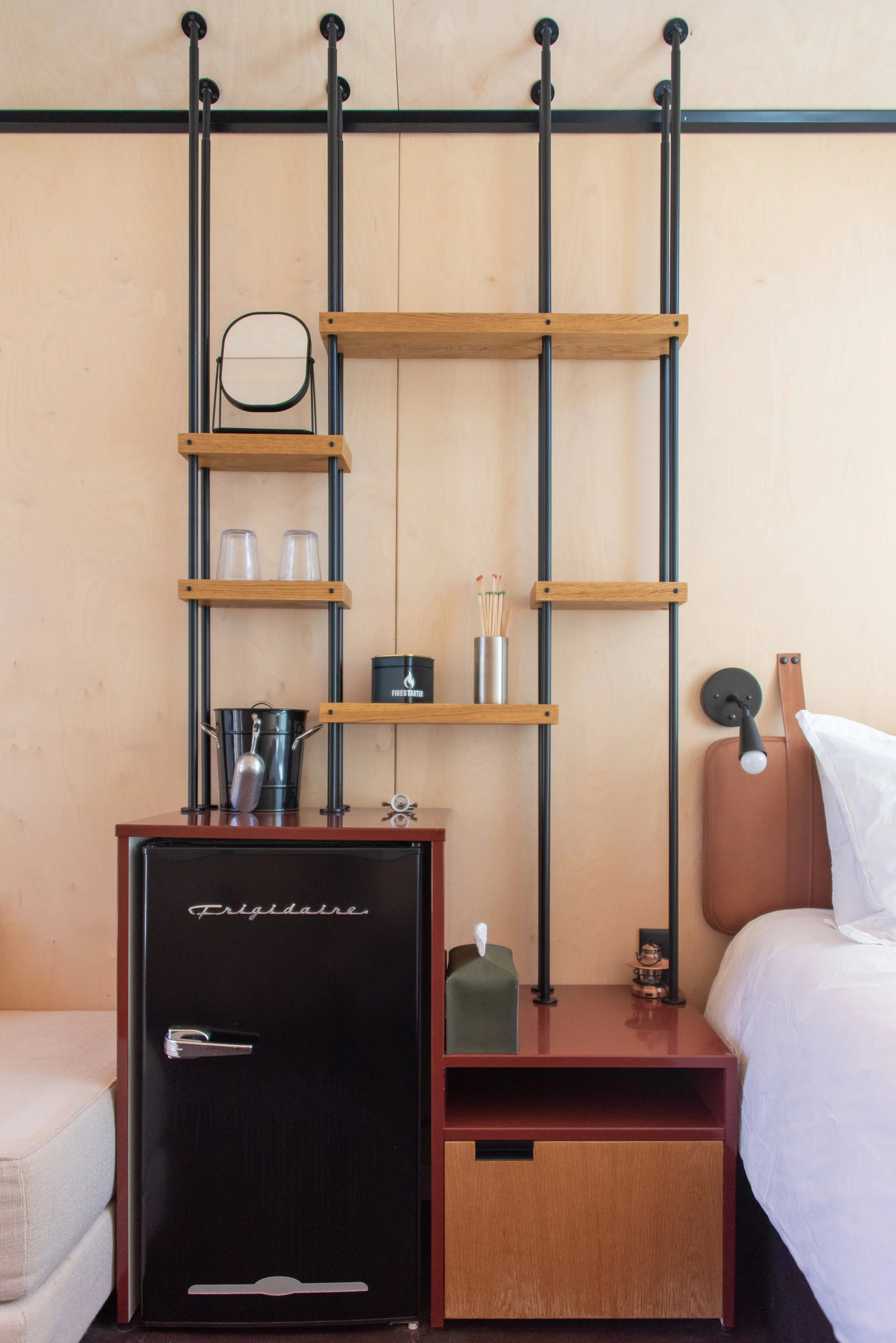 Tiny Cabin - Bedroom with a headboard, a nightstand with a green tissue box, and a shelving unit attached to the wall behind the bed. The unit holds glasses and small items, with a mini fridge below. Sleeps 2 people. 