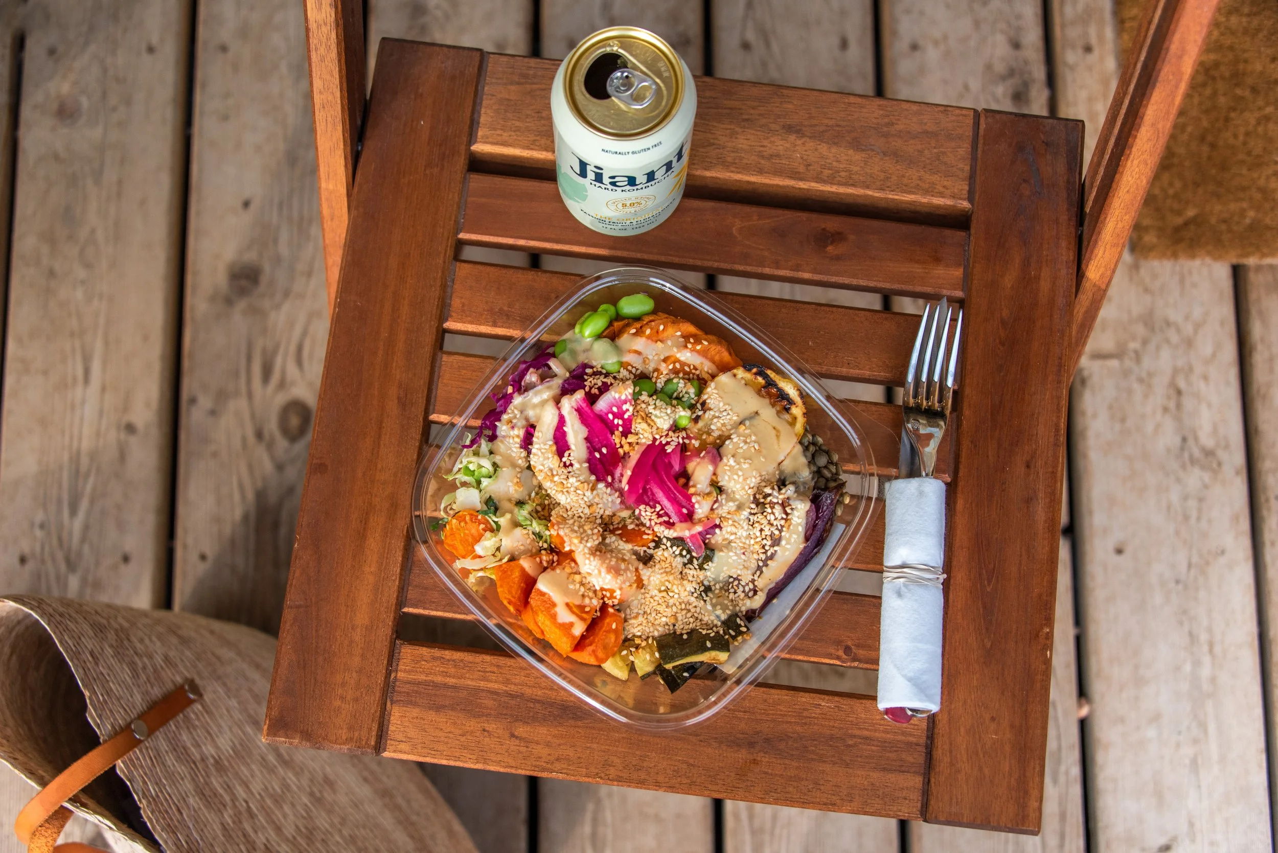 A wooden chair with a plastic container of colorful salad on it, a can of Jämt soda, and a fork wrapped in a napkin.