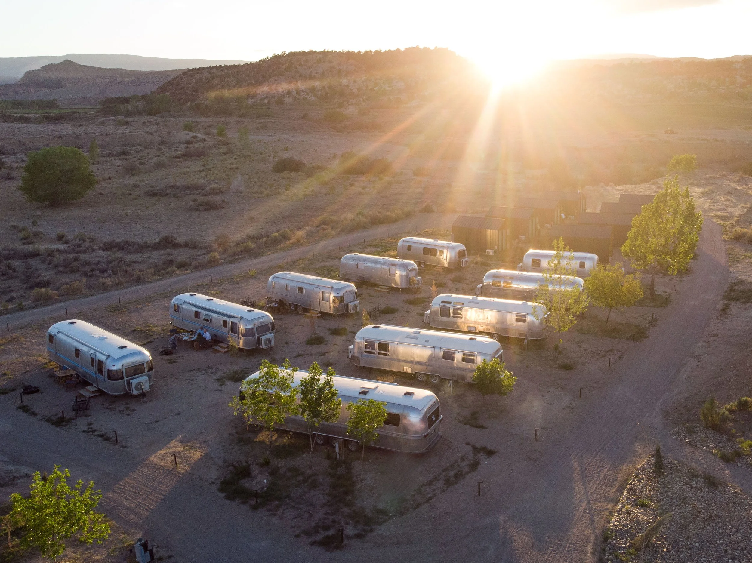 Aerial view of an outdoor hotel with multiple vintage Airstreams and tiny cabins among green trees, under a bright setting sun in a desert landscape with hills in the background.