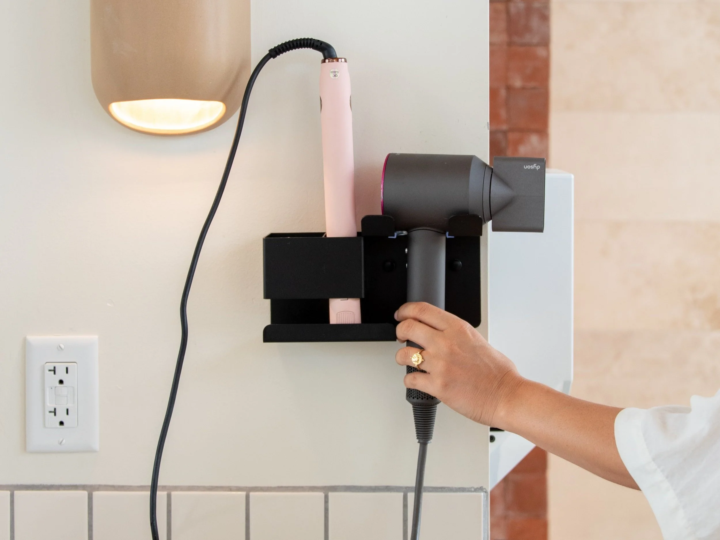 Spa-Quality Bathhouses - A person plugging a black Dyson hairdryer and pink straightener into a wall-mounted black holder on a kitchen or bathroom wall.