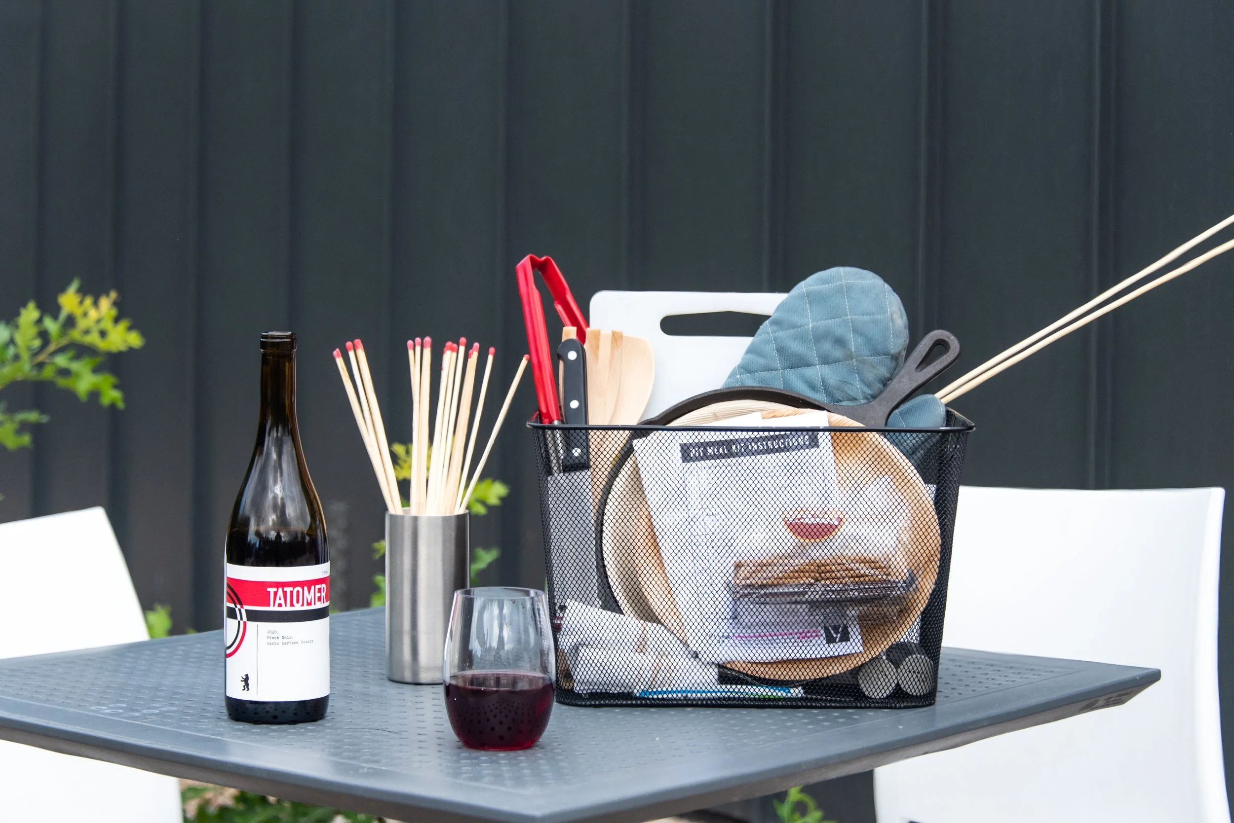 A table with a bottle of red wine, two glasses of red wine, a cup with chopsticks, and a metal basket filled with various kitchen utensils, including a red-handled saw, a white cutting board, a blue oven mitt, and some boxes. The background is a dark blue wall, and there is a small plant on the left side of the table.