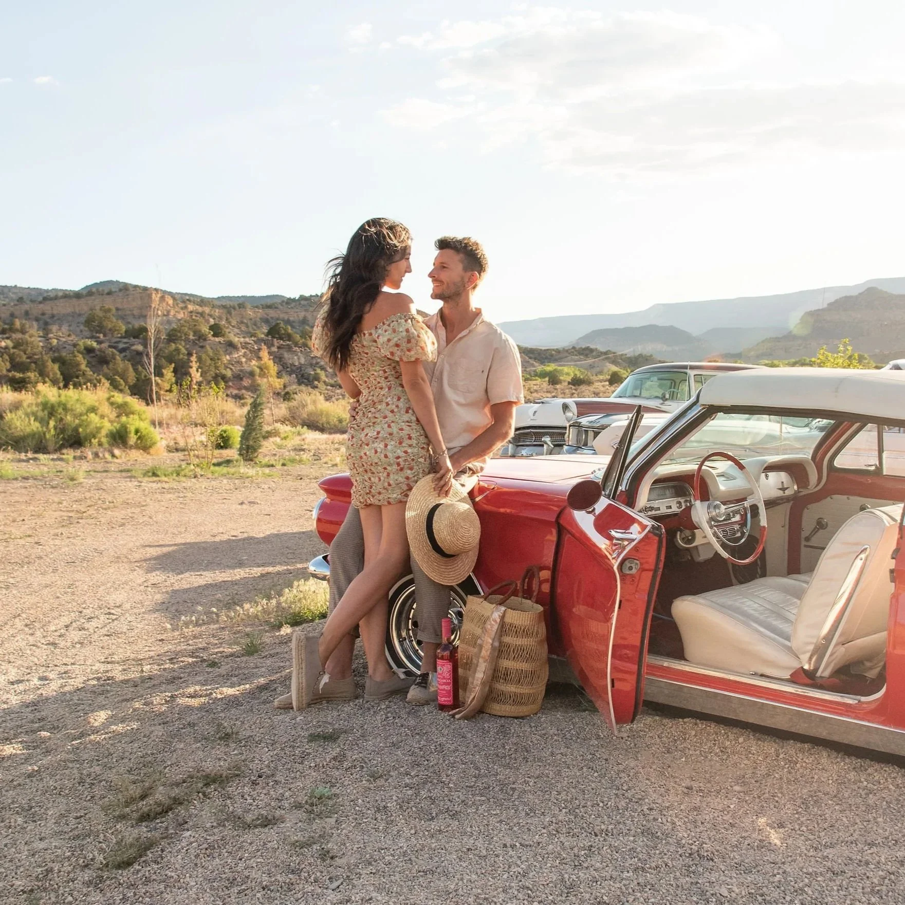 A man and woman standing close together next to a red vintage car in a desert landscape with mountains in the background. They are smiling at each other, holding a straw hat. The woman is wearing a floral dress, and the man is dressed in a light shirt and pants. There are baskets, a water bottle, and the car's interior visible, indicating a picnic or outing.